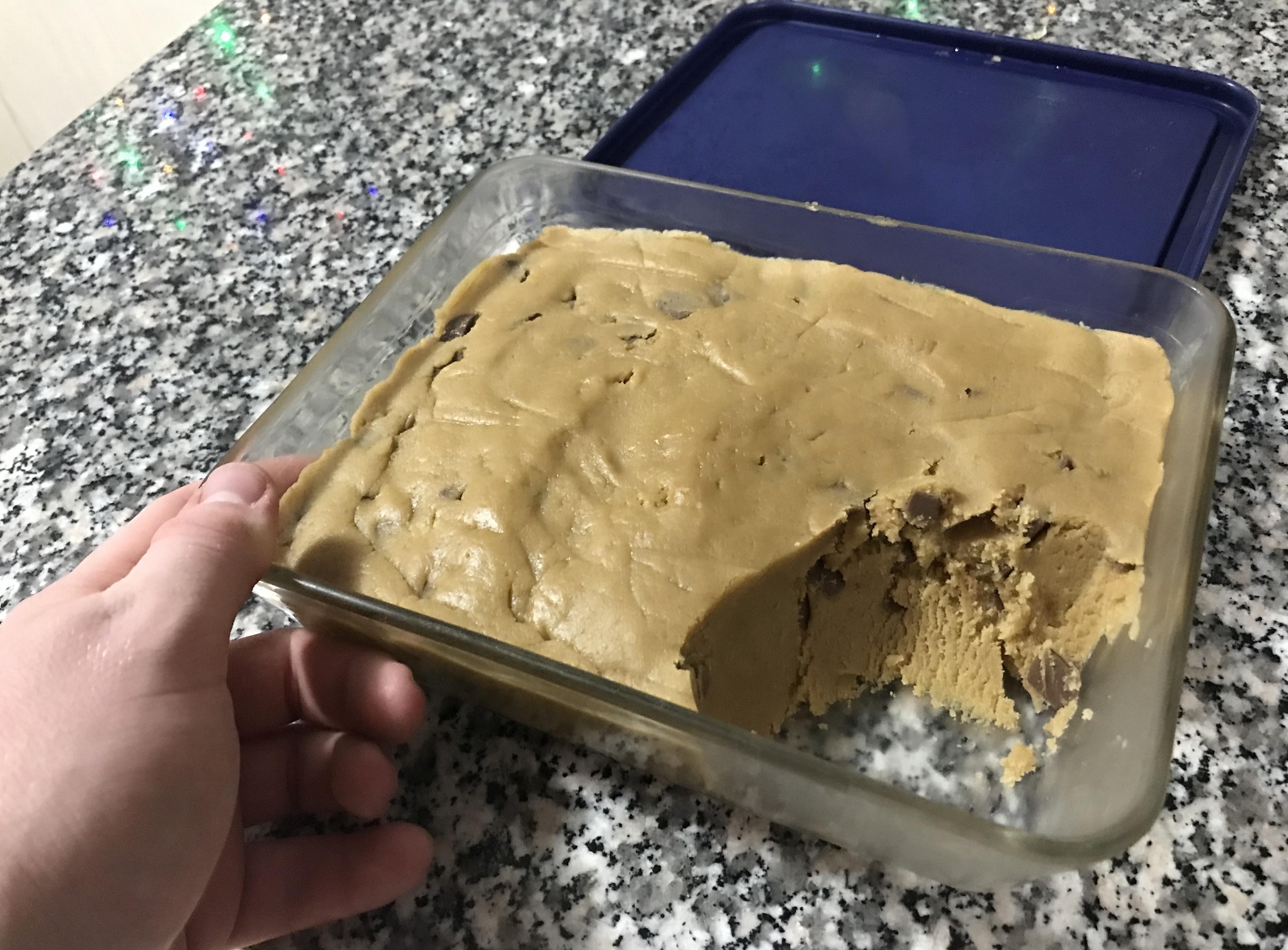 A hand holds a glass dish of chocolate chip cookie dough with a large bite taken out from one corner, sitting on a speckled countertop.