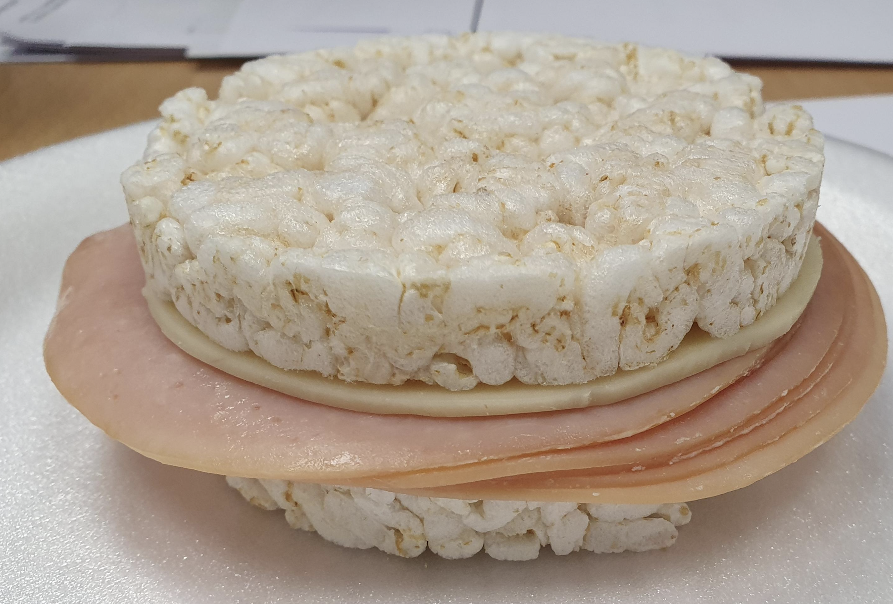 A sandwich made with two rice cakes, containing a slice of cheese and slices of ham, placed on a white surface.