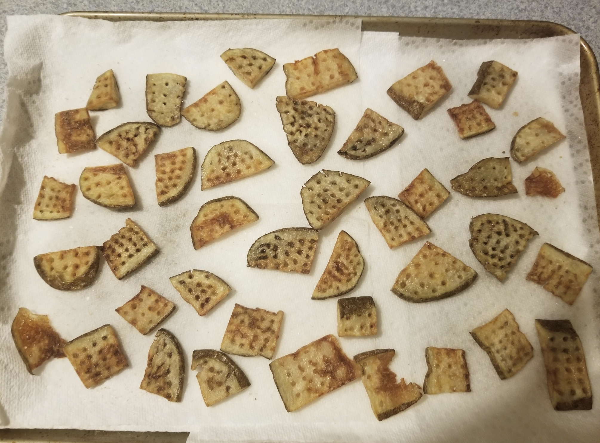 Baked potato slices of various shapes and sizes are spread out on a paper towel-lined baking sheet, cooling after being cooked.
