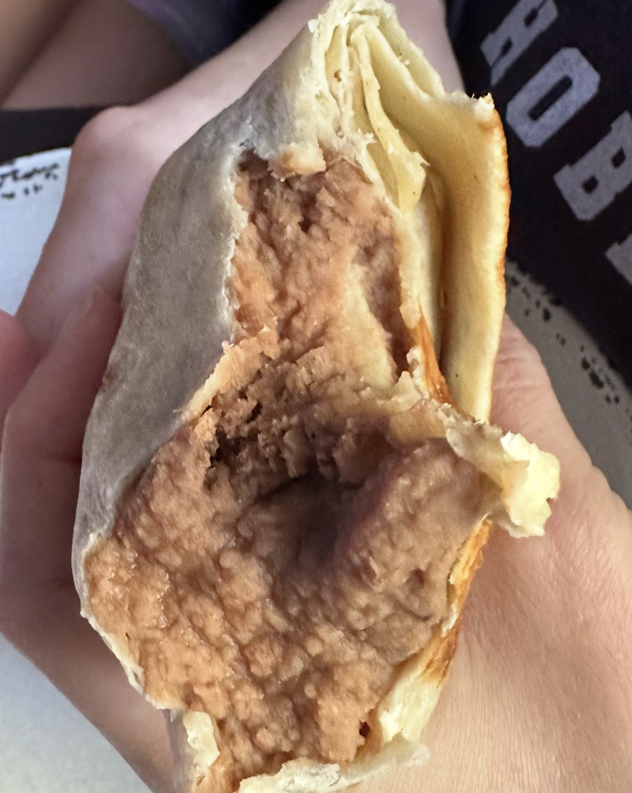 A hand holds a burrito with a flour tortilla, revealing a filling of refried beans. A person is in the background, partially visible, wearing a dark shirt.