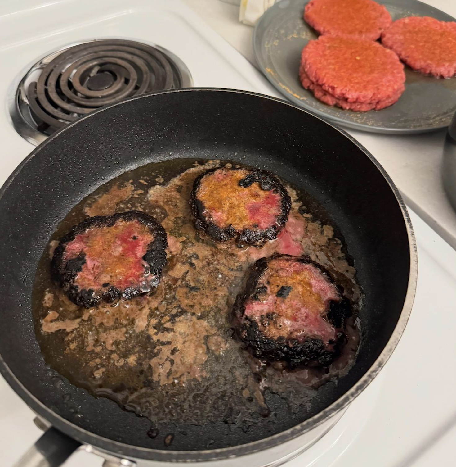 Three burger patties are burning in a frying pan on a stove, with pink raw patties on a plate nearby. The cooked patties have charred edges and are sitting in bubbling oil.
