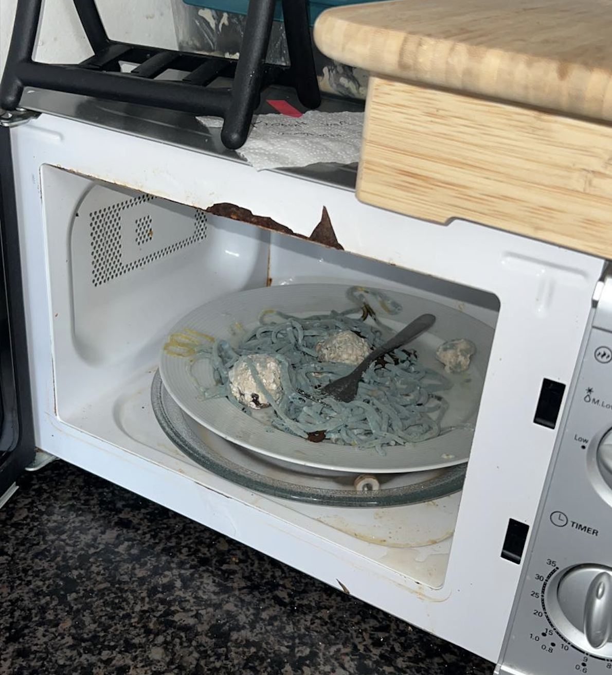 Open microwave with blue noodles and a fork on the glass plate inside. The microwave’s interior shows rust and damage, and it sits on a dark countertop under a wooden cutting board and a black kitchen rack.