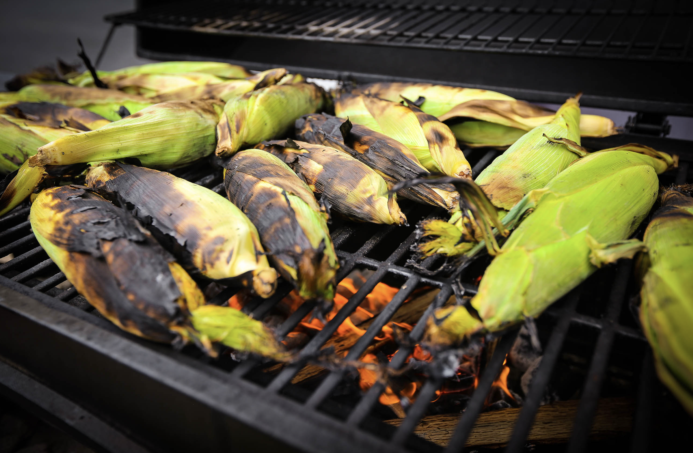 Several ears of corn in husks are being grilled over an open flame, with some husks charred and some still green. The grill grates and orange fire are visible beneath the corn.