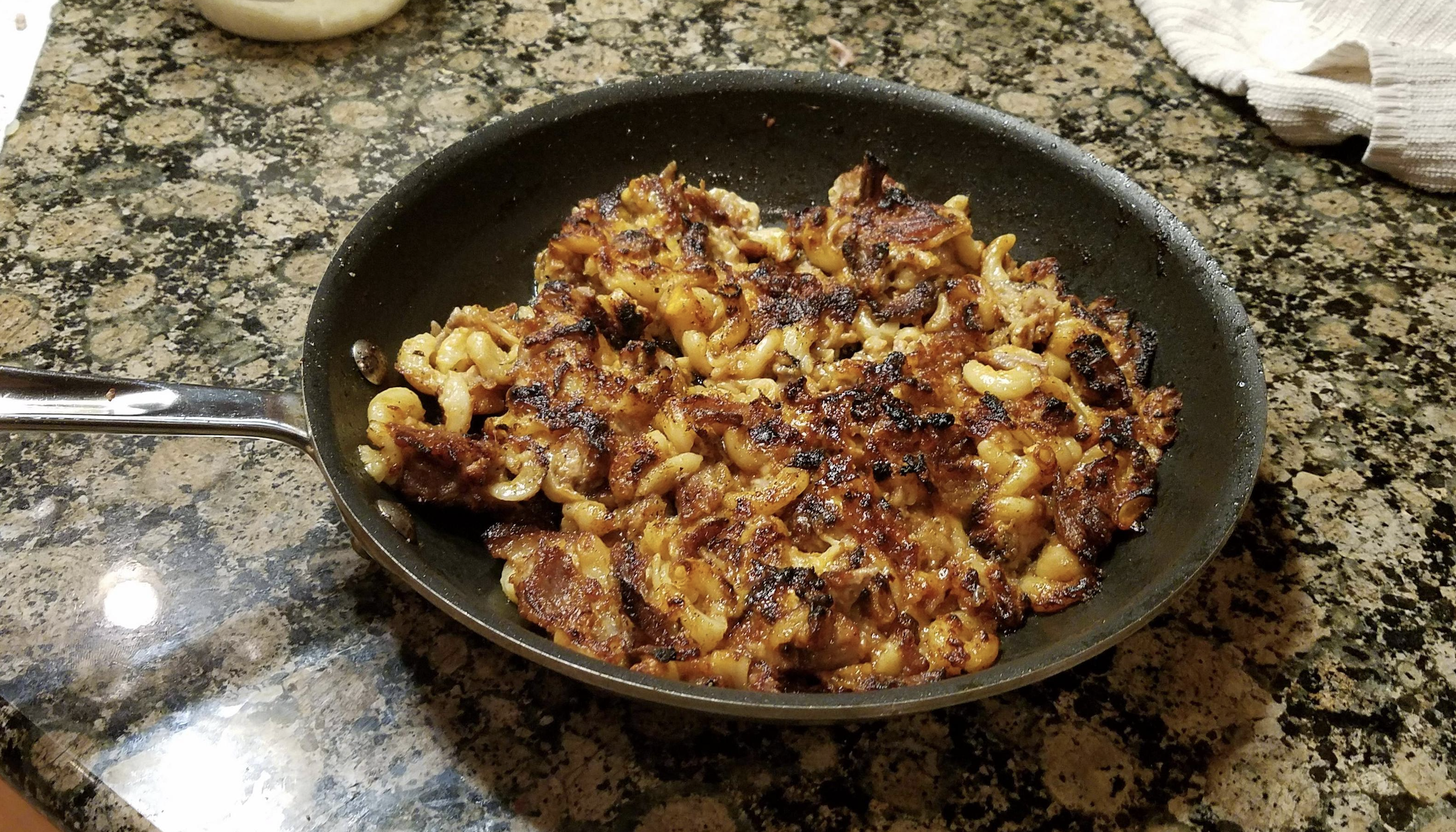 A frying pan on a granite countertop holds crispy, browned macaroni and cheese, showing a golden, slightly charred crust. A white towel is in the upper right corner.