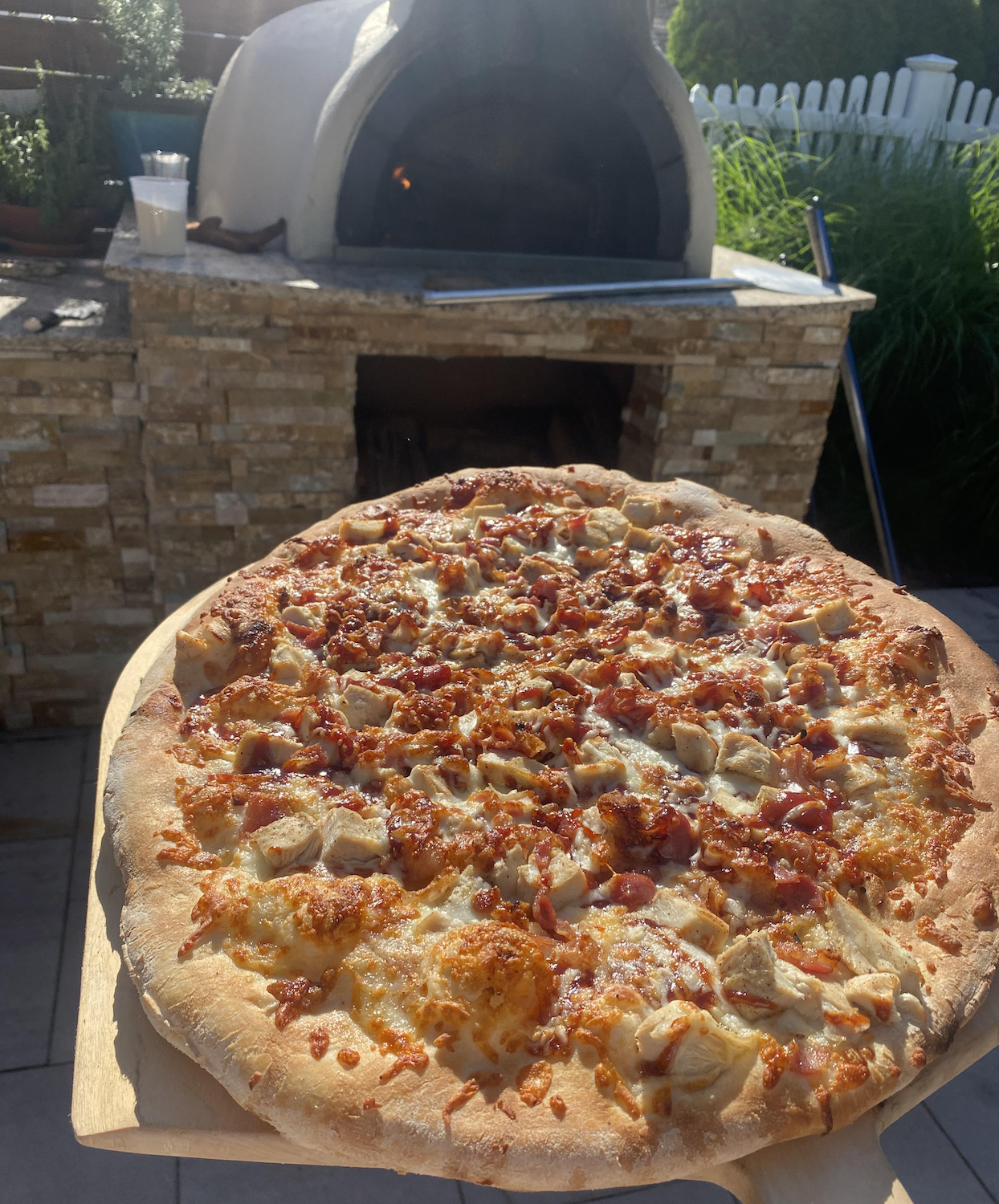 A freshly baked pizza with chicken and bacon is held in front of a stone wood-fired pizza oven outdoors on a sunny day.