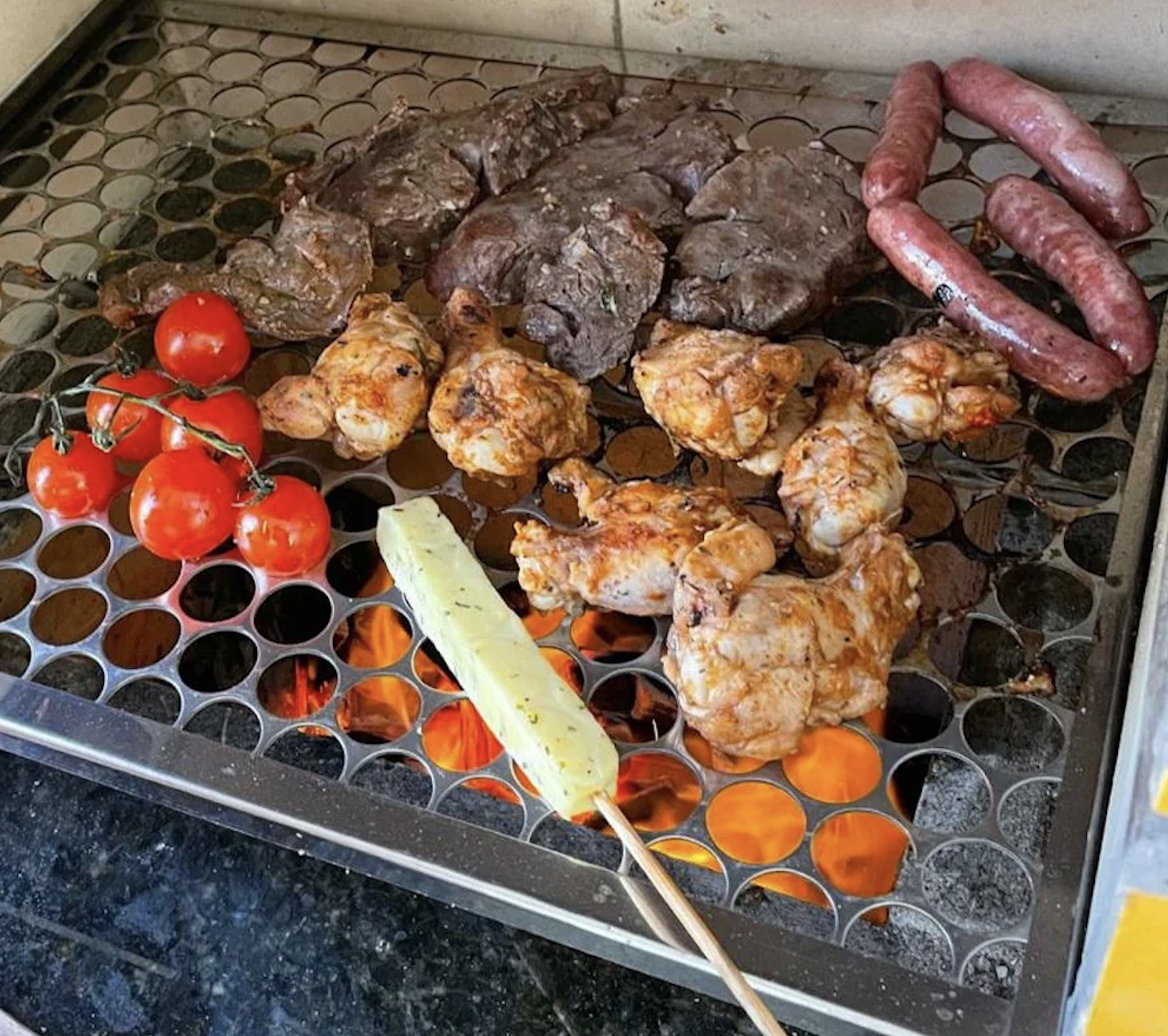 Various meats, sausages, skewered chicken, a cheese stick, and a bunch of cherry tomatoes are grilling over an open flame on a metal grate.