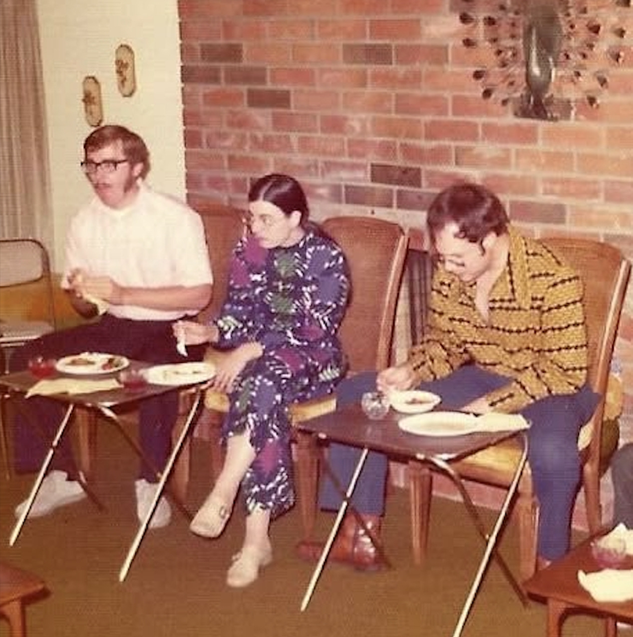 Three people sit on wicker chairs eating food from trays in a vintage living room with brick wall, peacock decor, and carpet. They appear to be at a casual gathering in the 1970s.