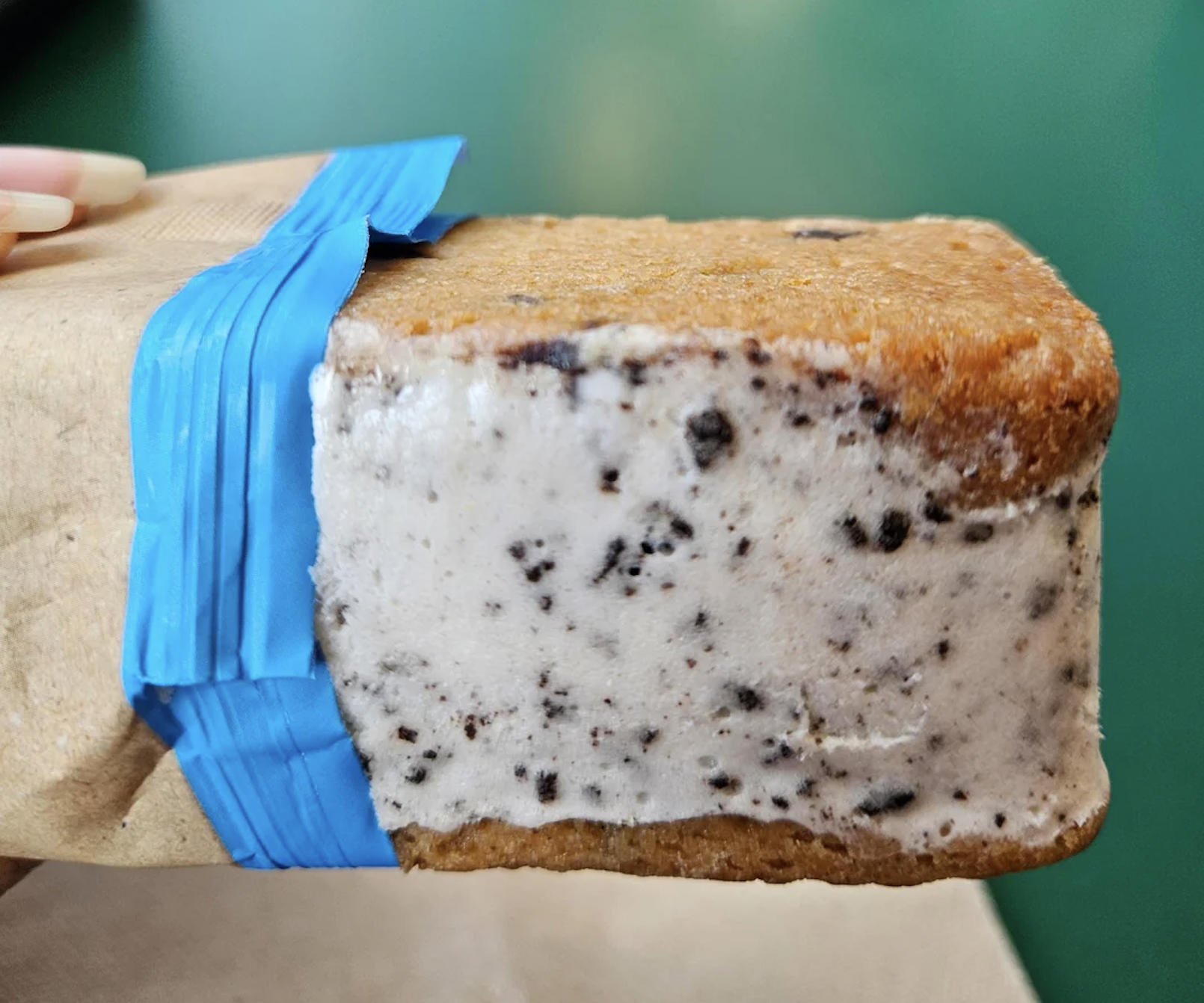 A close-up of an ice cream sandwich with cookies and cream filling between two brown cookies, partially wrapped in brown paper and secured with blue tape, against a green background.