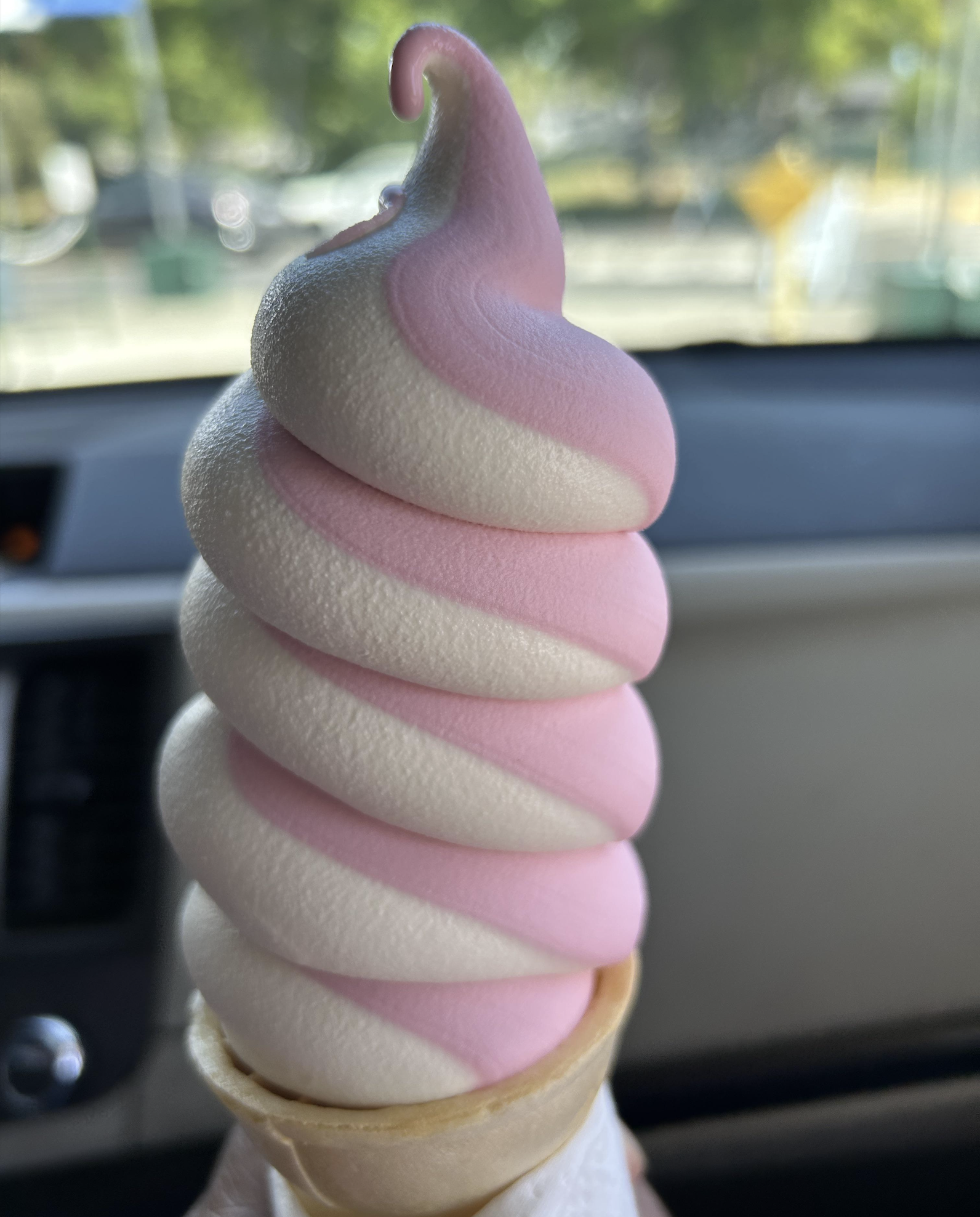 A close-up of a pink and white swirled soft serve ice cream in a cone, held inside a car with blurred trees and sunlight visible through the windows in the background.