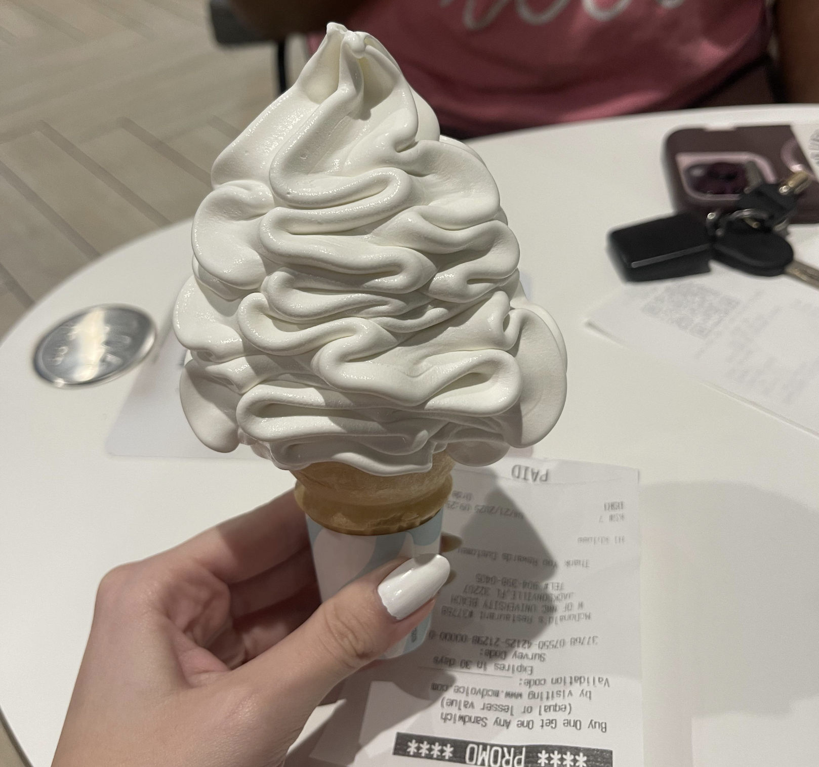 A hand holding a soft-serve vanilla ice cream cone with a receipt, on a white table with car keys and a phone in the background.
