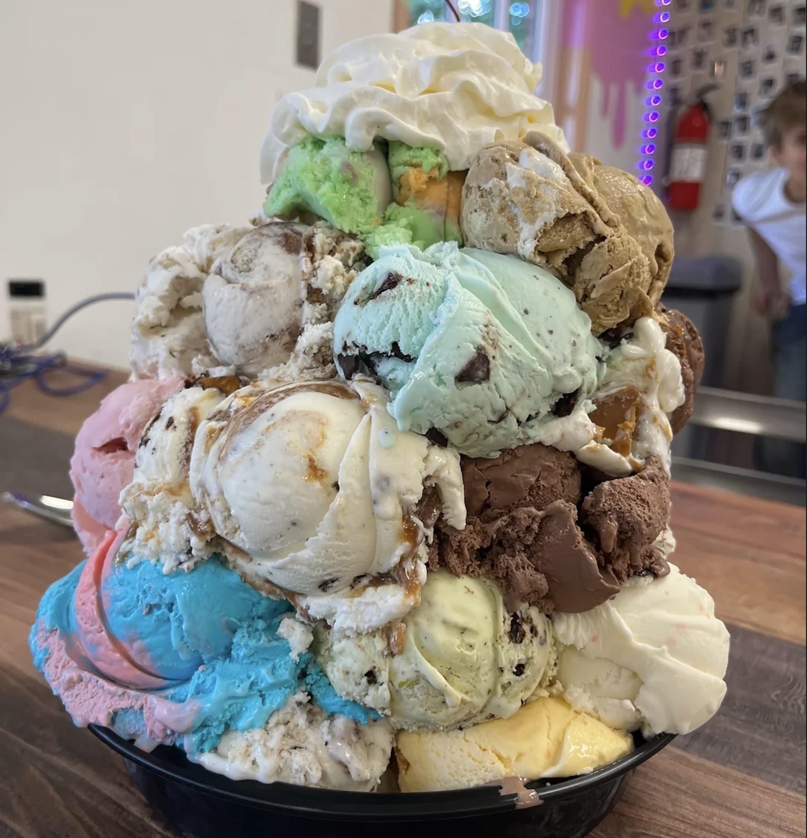 A large bowl piled high with many colorful scoops of ice cream, topped with whipped cream, sits on a wooden counter. A child stands in the background near a wall with photos and neon lights.
