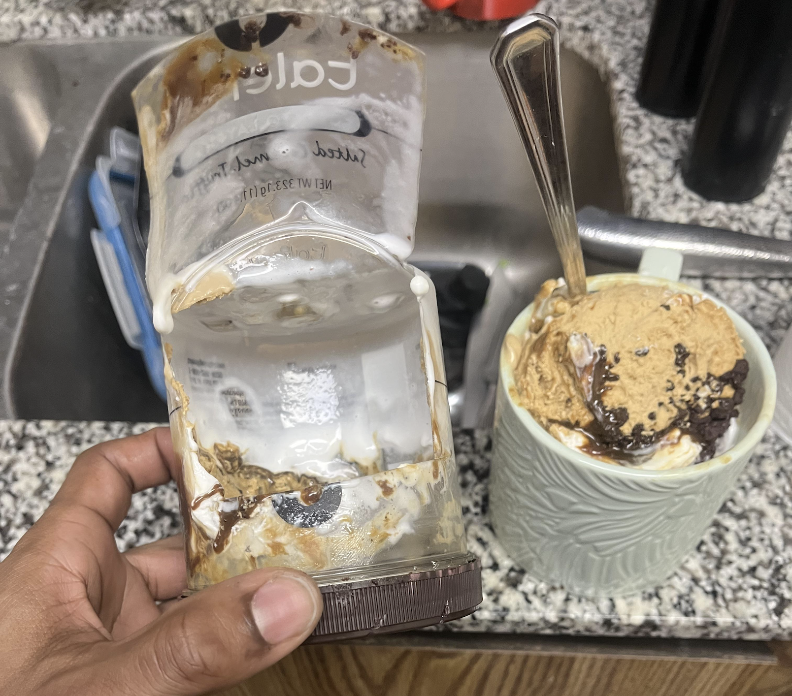 A hand holds an empty, messy container of caramel sauce over a granite countertop near a sink. Next to it is a mug filled with ice cream topped with caramel and chocolate syrup and a spoon inside.