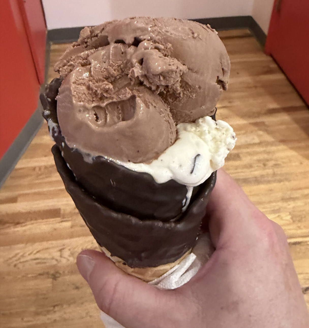 A hand holds a chocolate-dipped ice cream cone with scoops of chocolate and vanilla ice cream, set against a background of wooden flooring and red doors.