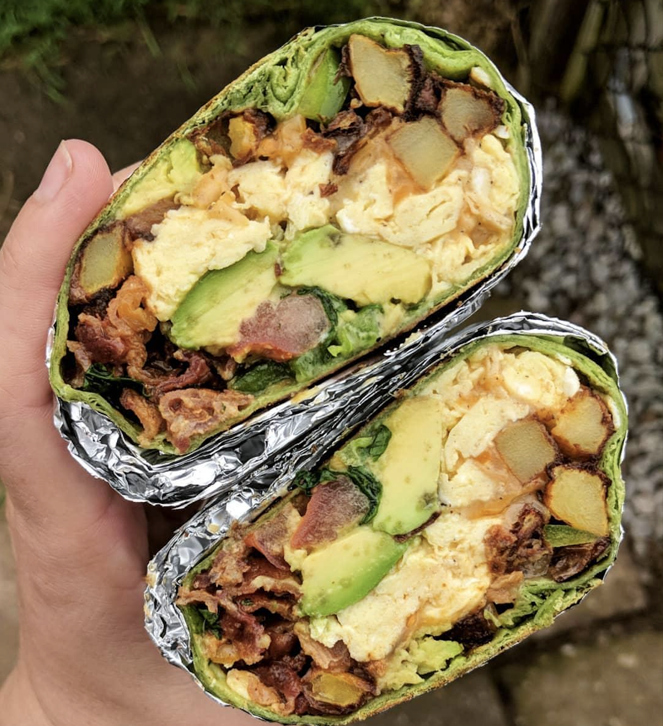 A hand holds a halved breakfast burrito filled with scrambled eggs, avocado slices, potatoes, bacon, and veggies, wrapped in foil and a green tortilla.