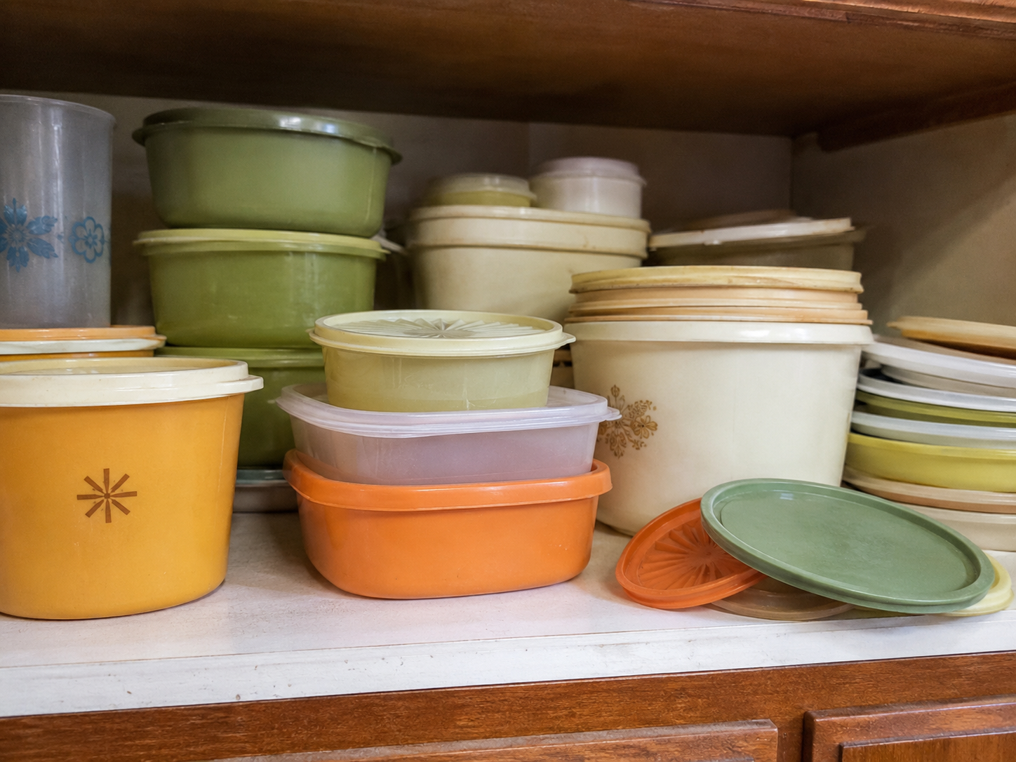 An assortment of vintage plastic food storage containers, mostly in green, yellow, orange, and beige colors reminiscent of those popular in the 80s, is stacked and arranged on a white kitchen shelf.