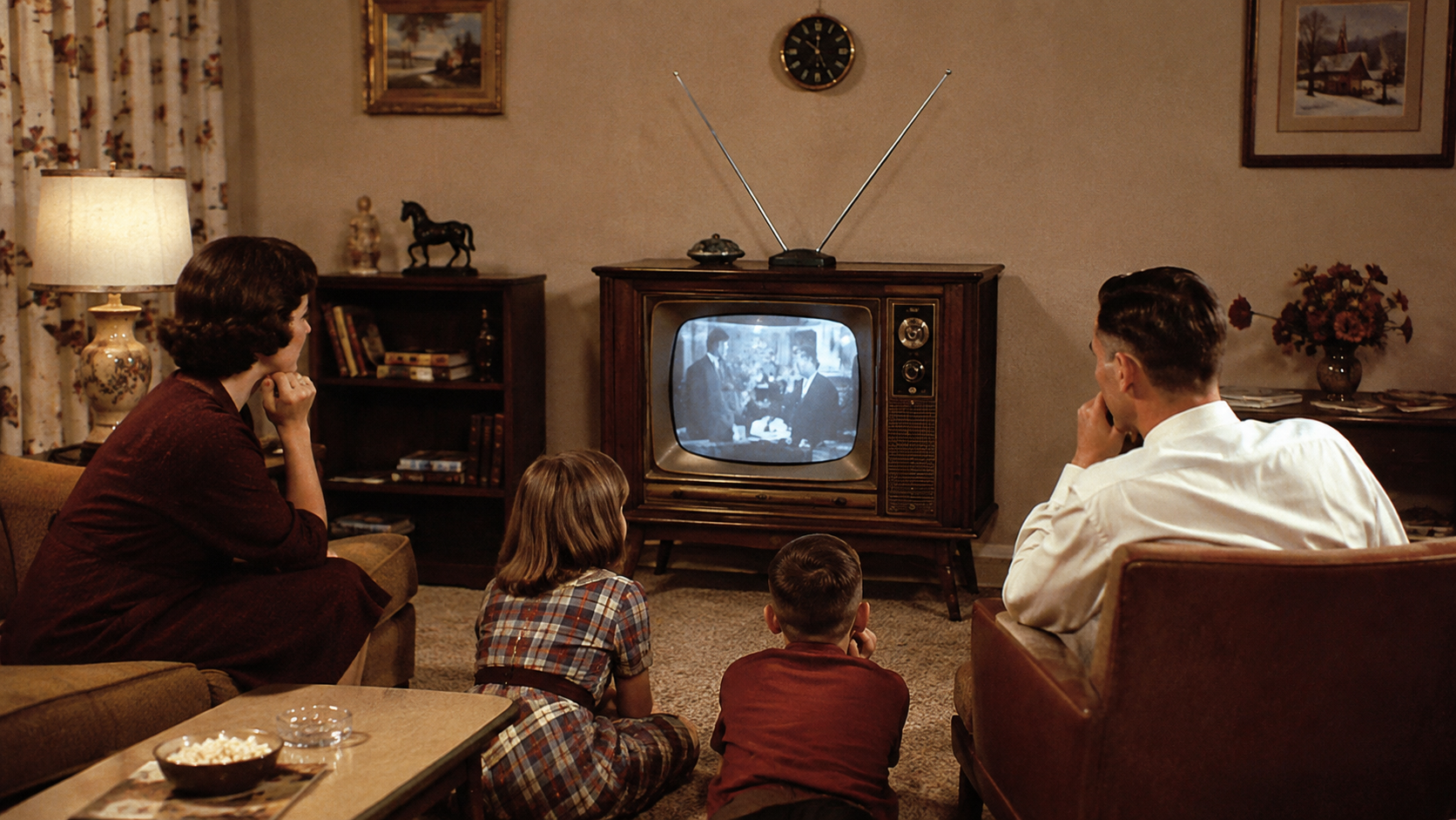 A family of four sits in a cozy living room, watching a black-and-white program on a vintage television with antennae. The room is warmly lit and decorated in a classic mid-century style, perfectly capturing the charm of Homes In The 60s.