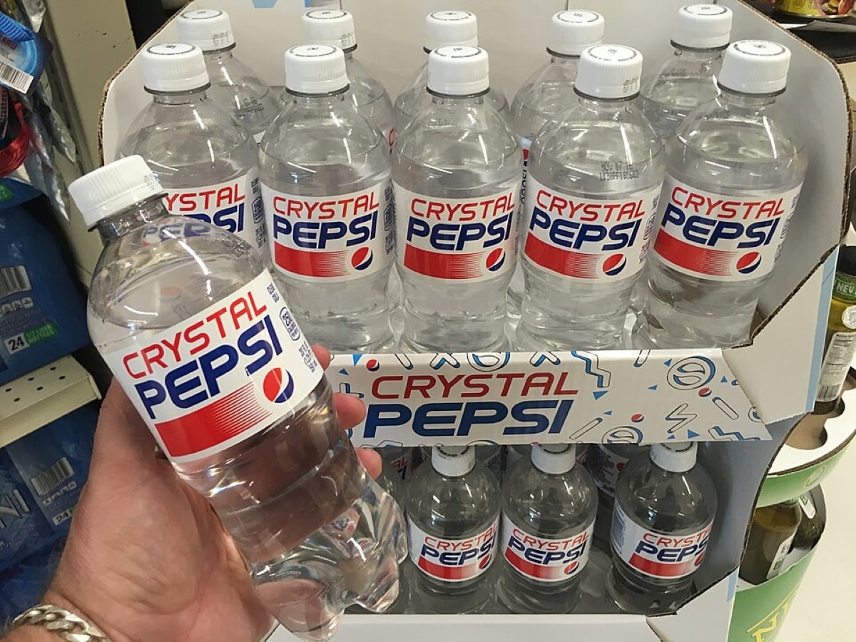 A hand holds a bottle of Crystal Pepsi, one of the iconic soda pops, in front of a display filled with more Crystal Pepsi bottles. The bottles contain clear soda and have a red, white, and blue label.