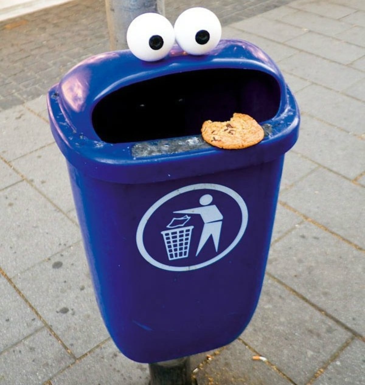 A blue trash bin with googly eyes and a cookie on its rim, resembling Cookie Monster from Sesame Street, stands on a sidewalk.