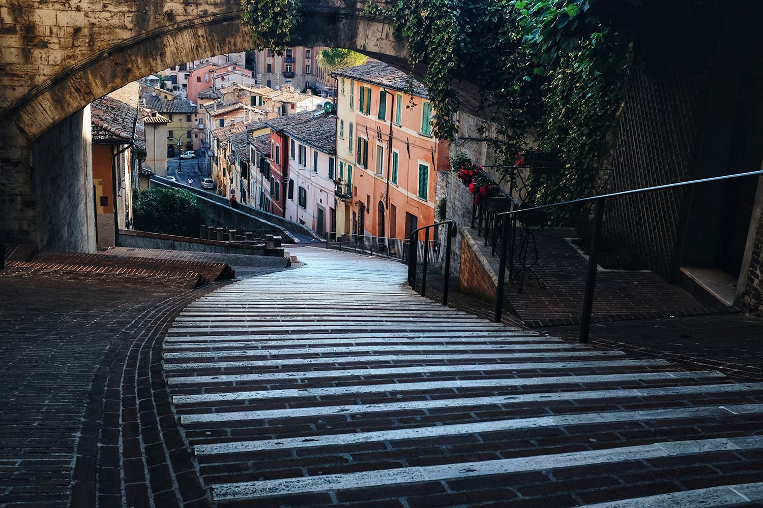 A curved stone staircase leads downhill under an arched bridge, with colorful buildings, potted plants, and ivy visible along the street in a charming European town.