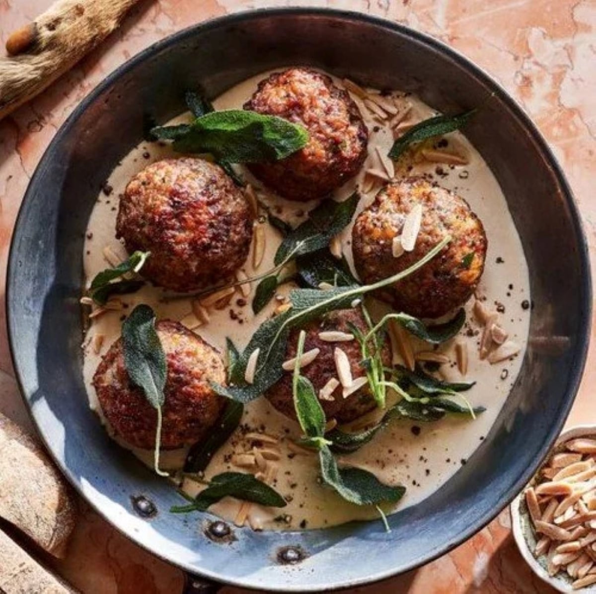 Five browned meatballs sit in a creamy sauce in a metal pan, topped with fried sage leaves and sliced almonds, on a marble surface.