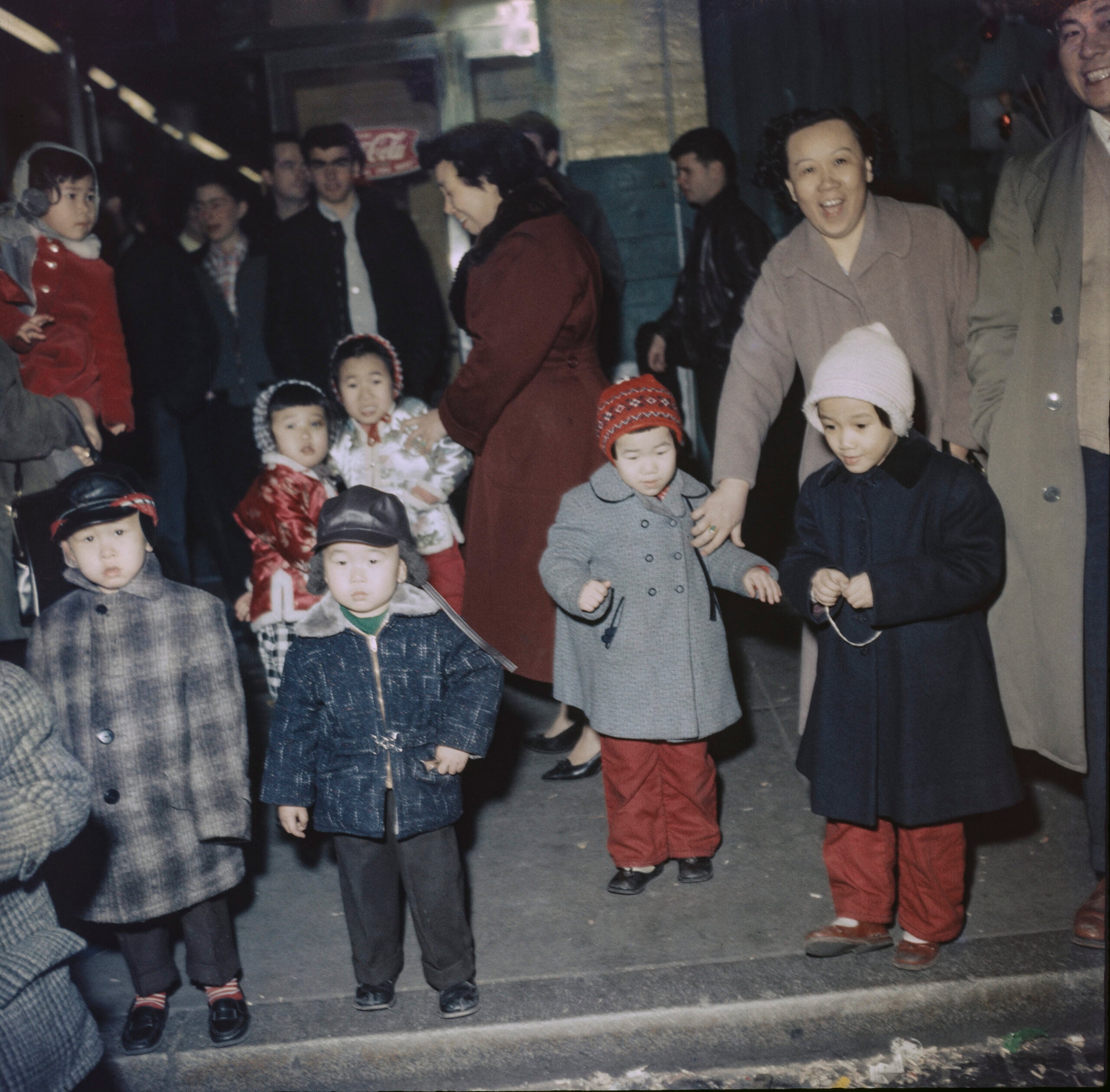 A group of children in coats and hats stand on a city street, with adults nearby. The scene appears festive, with people smiling and engaged in conversation.