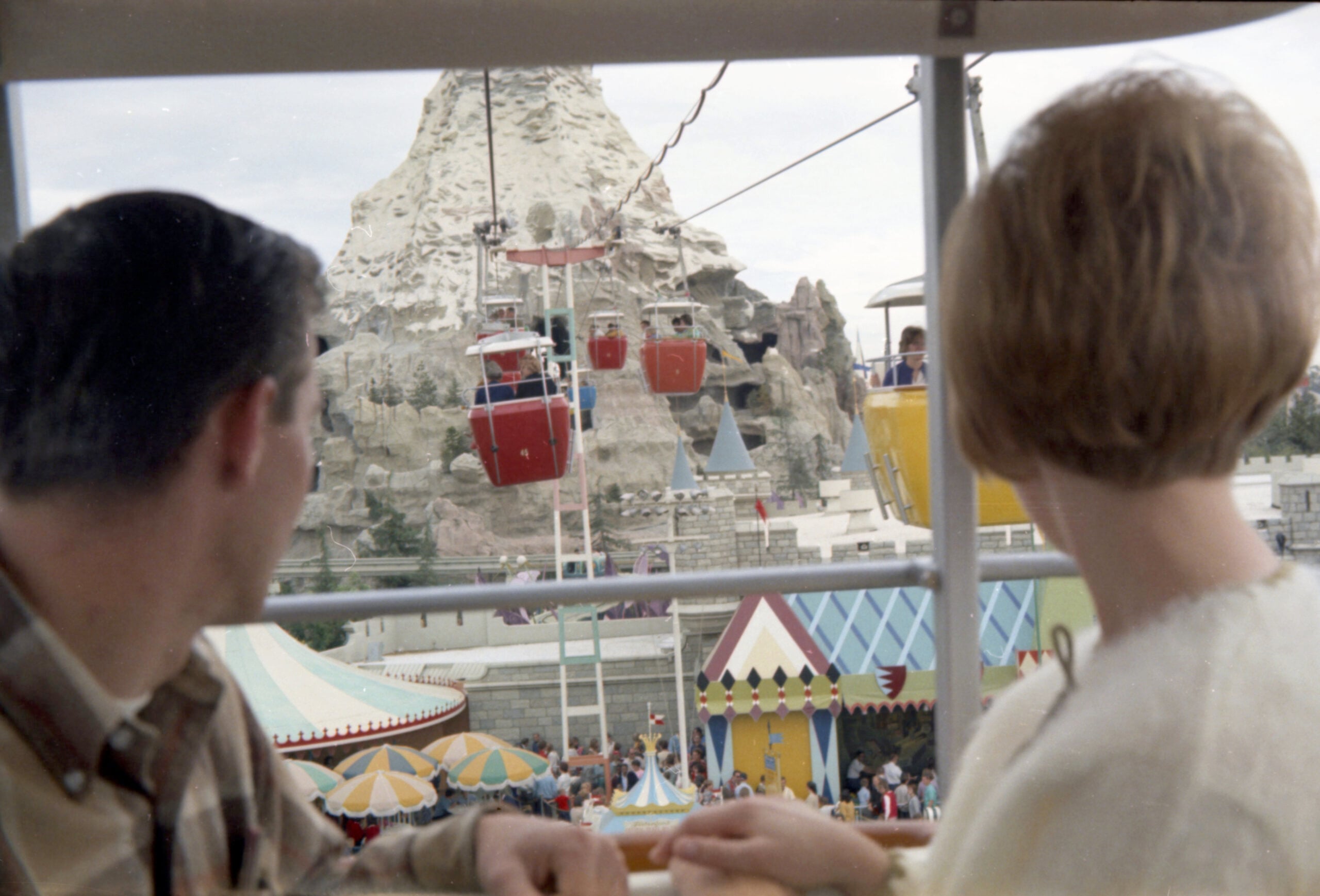 A man and woman look out from a ride at colorful gondolas and a mountain at an amusement park, with crowds, tents, and festive scenery below.