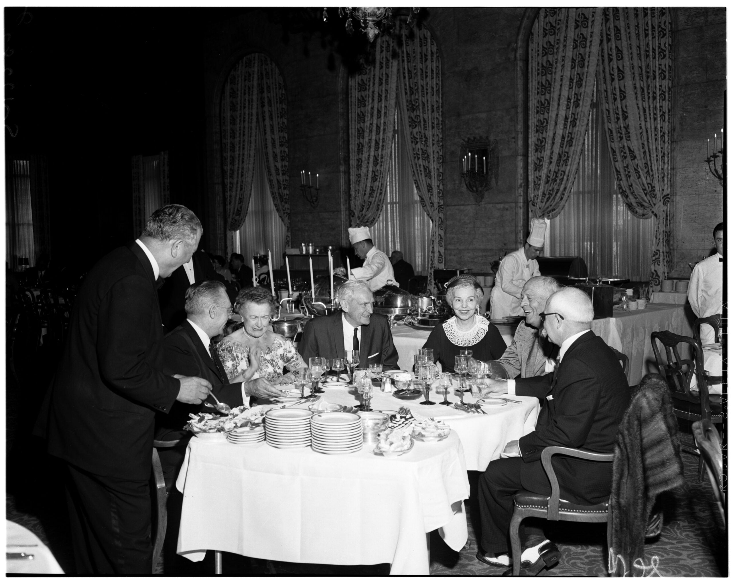 A group of well-dressed people sit around a table at a formal dinner in an elegant dining room, with chefs serving food in the background.