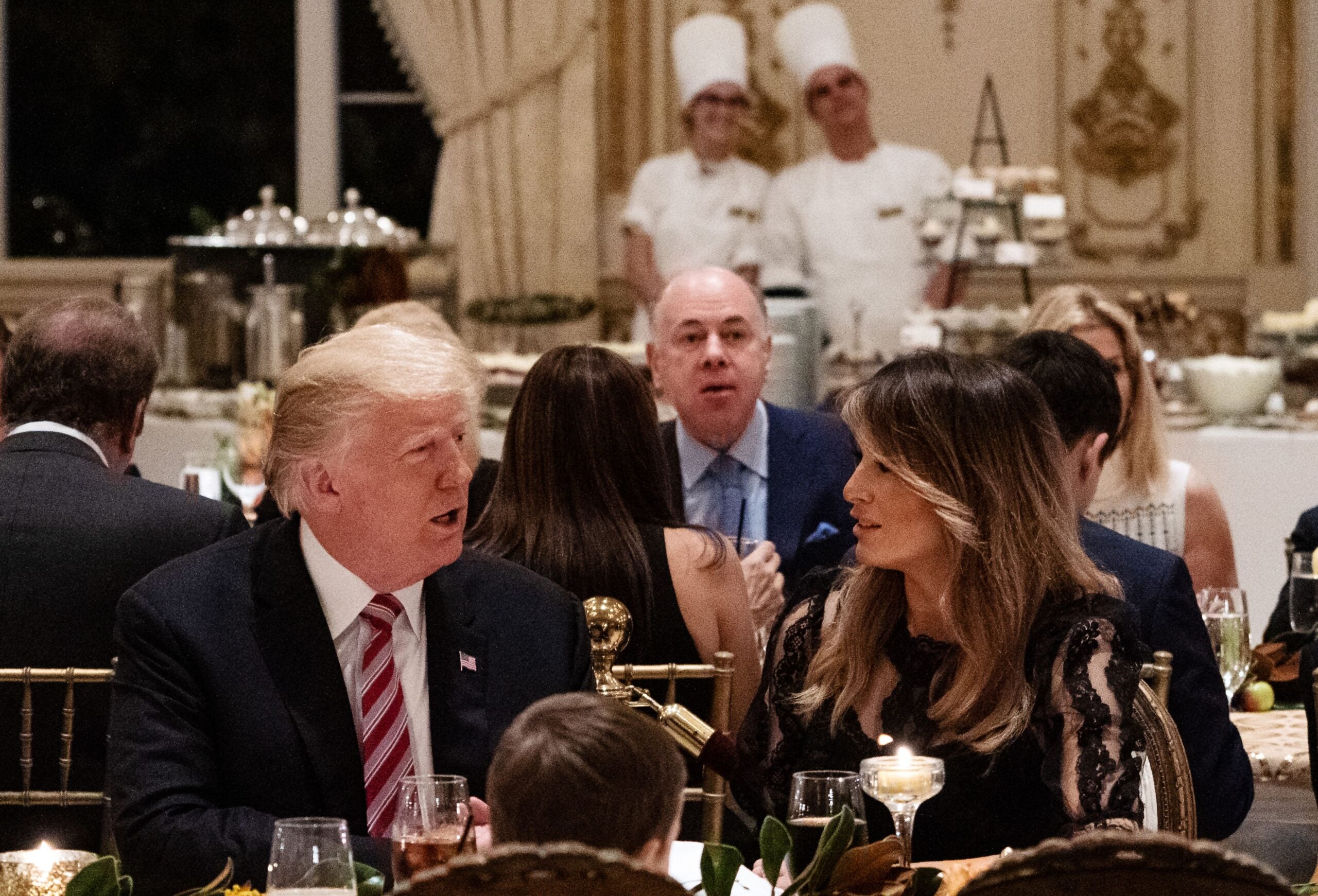A man and woman sit at a formal dinner table, speaking to each other. Other guests and two chefs in white uniforms are seen in the background in an ornate dining room.