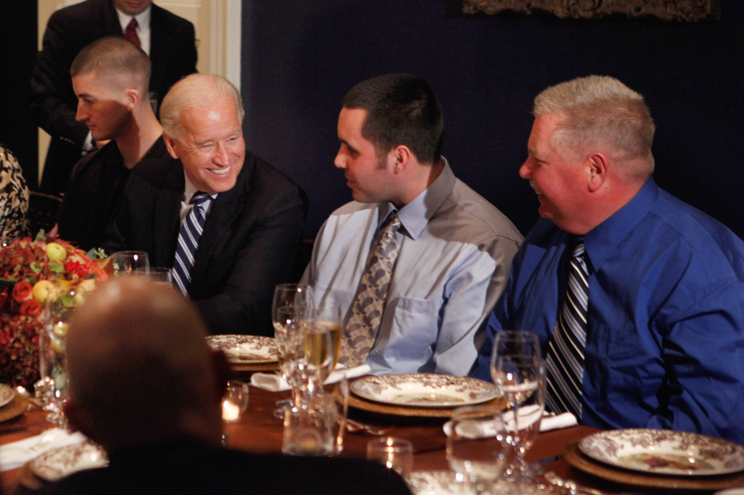 Four men in dress shirts and ties sit together at an elegantly set dinner table, smiling and conversing. A floral centerpiece and drinks are on the table. The atmosphere appears formal and friendly.