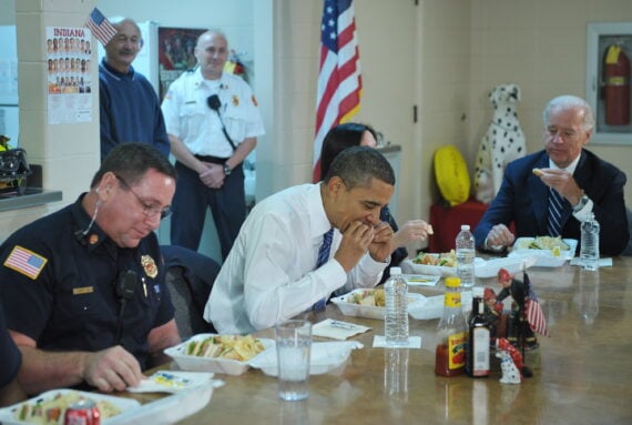 Three men, including two in uniform and one in a suit, eat sandwiches at a table with American flags. Two other men stand in the background, one in uniform. An American flag and a Dalmatian statue are visible in the room.