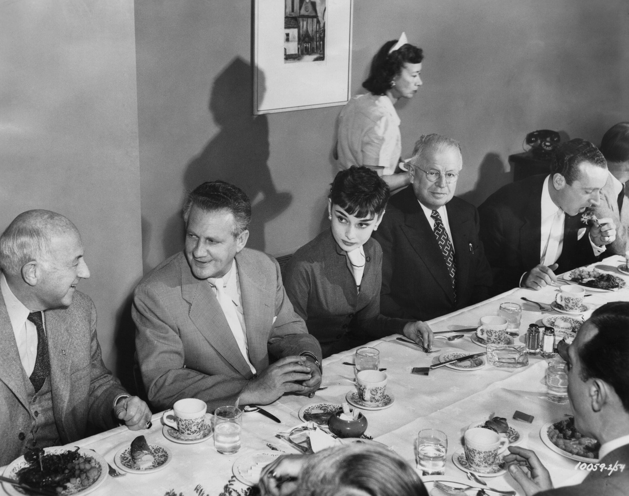 A group of people sit at a long dining table, dressed in formal attire, engaging in conversation. A woman in a uniform serves food in the background. The scene appears to be from a black-and-white film or mid-20th-century event.