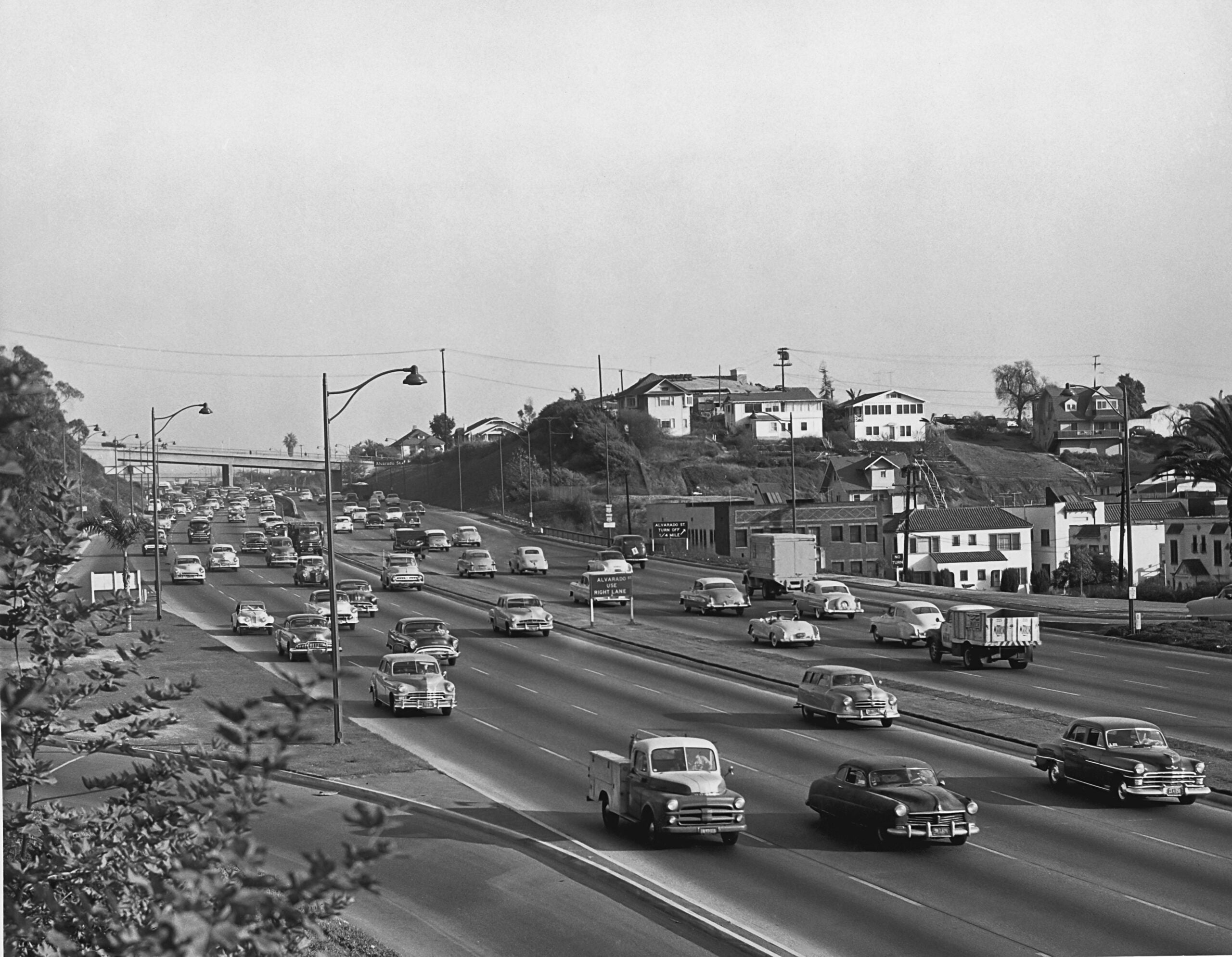 Black and white photo of a busy freeway with many 1950s cars and trucks driving in both directions. Houses and trees are visible on a nearby hill. Streetlights and a bridge cross above the road.