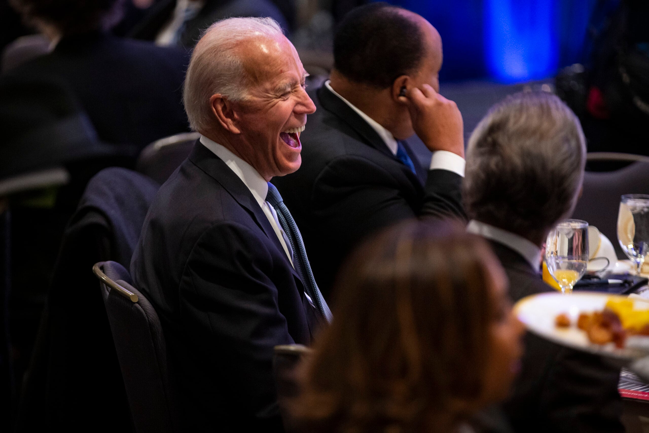 A man in a suit sits at a formal event, laughing heartily. Other attendees are seated nearby, and plates of food and drinks are on the tables.