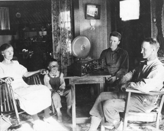 A family of four sits in a modest living room, gathered around a radio on a small table. Two adults and two children appear relaxed and attentive, listening together. The decor is simple and rustic.