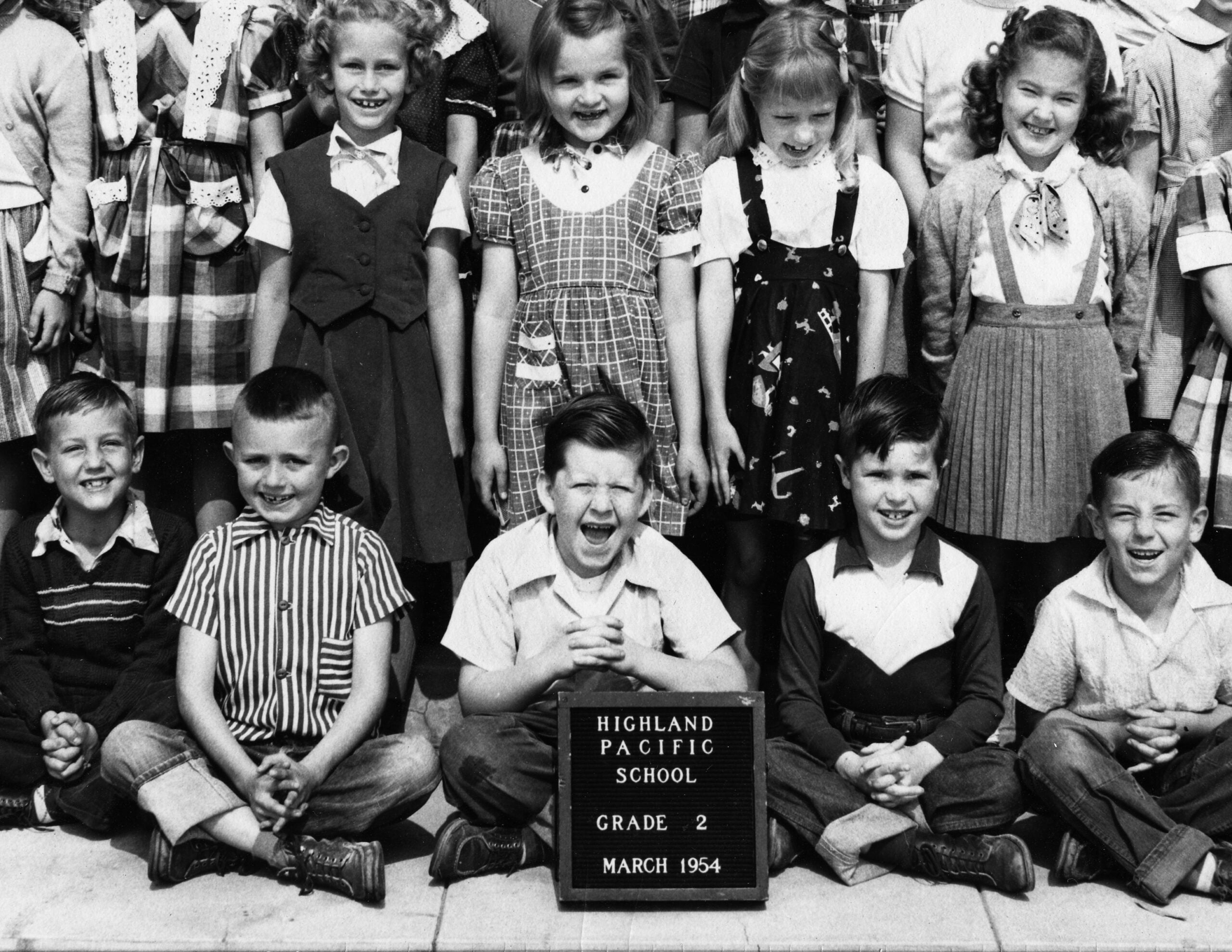 A black-and-white photo of children in two rows, with boys sitting and girls standing behind. A sign in front reads: “Highland Pacific School, Grade 2, March 1954.”