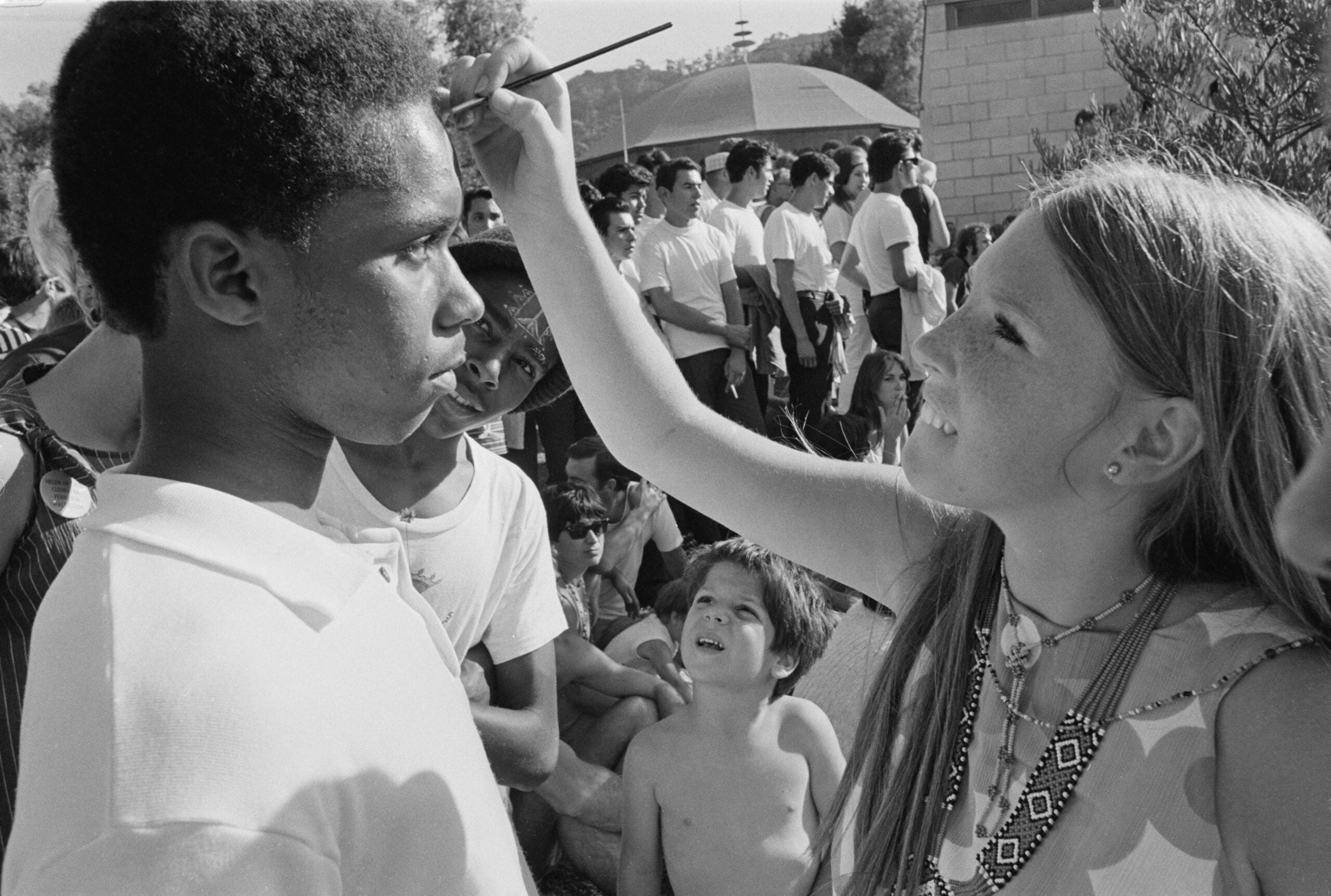 A young woman smiles while painting the forehead of a young man outdoors. A crowd of people, including children, watch in the background on a sunny day.