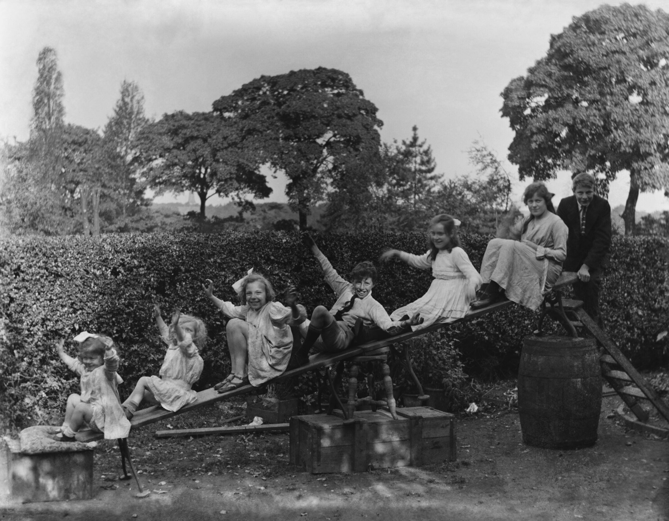 Seven children in old-fashioned clothes play on a homemade seesaw made from a plank and barrels outdoors, with hedges and trees in the background. Everyone looks happy and playful.