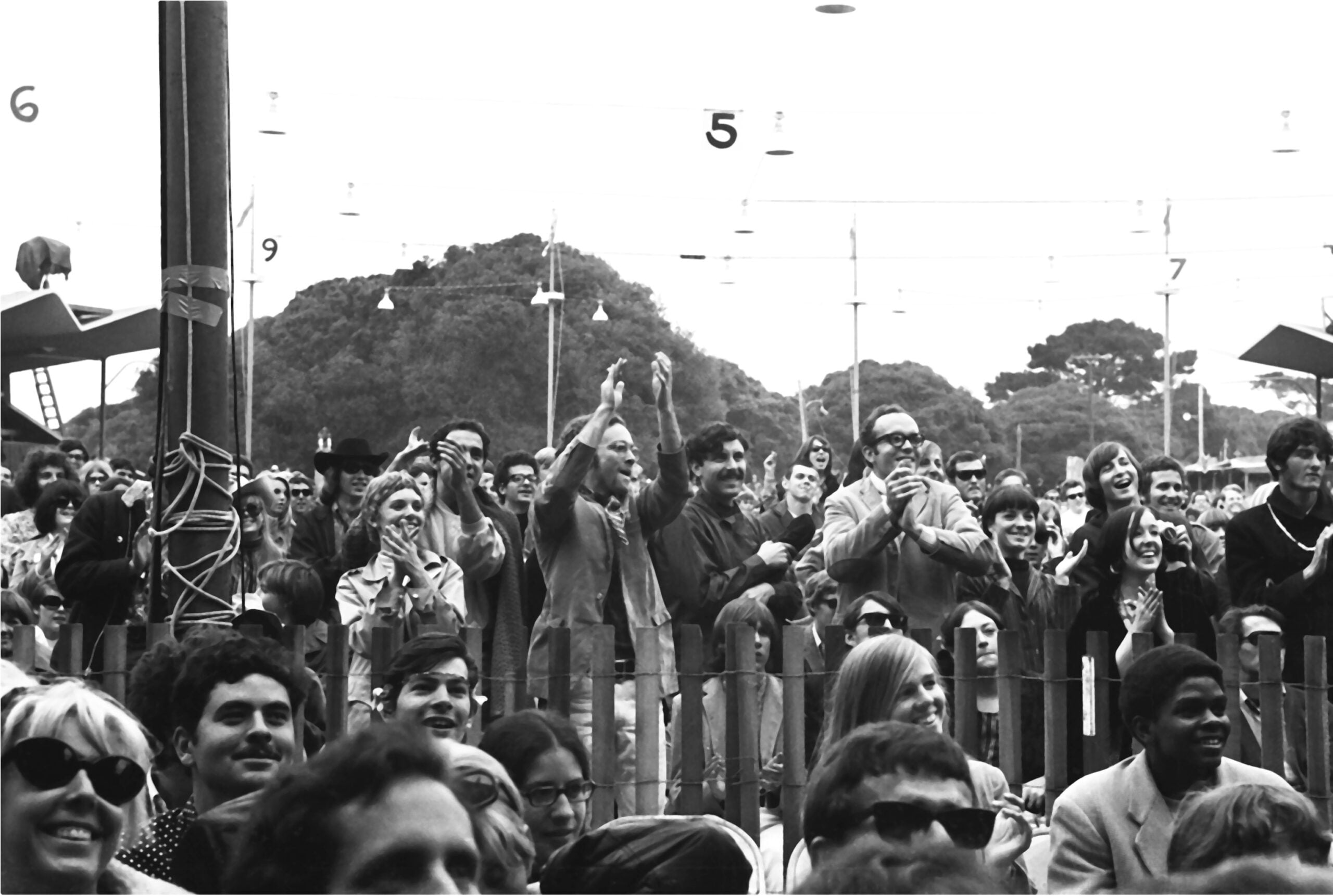 A large, diverse crowd of people outdoors, many smiling, clapping, and cheering, stand behind a short fence with numbered signs and trees visible in the background.