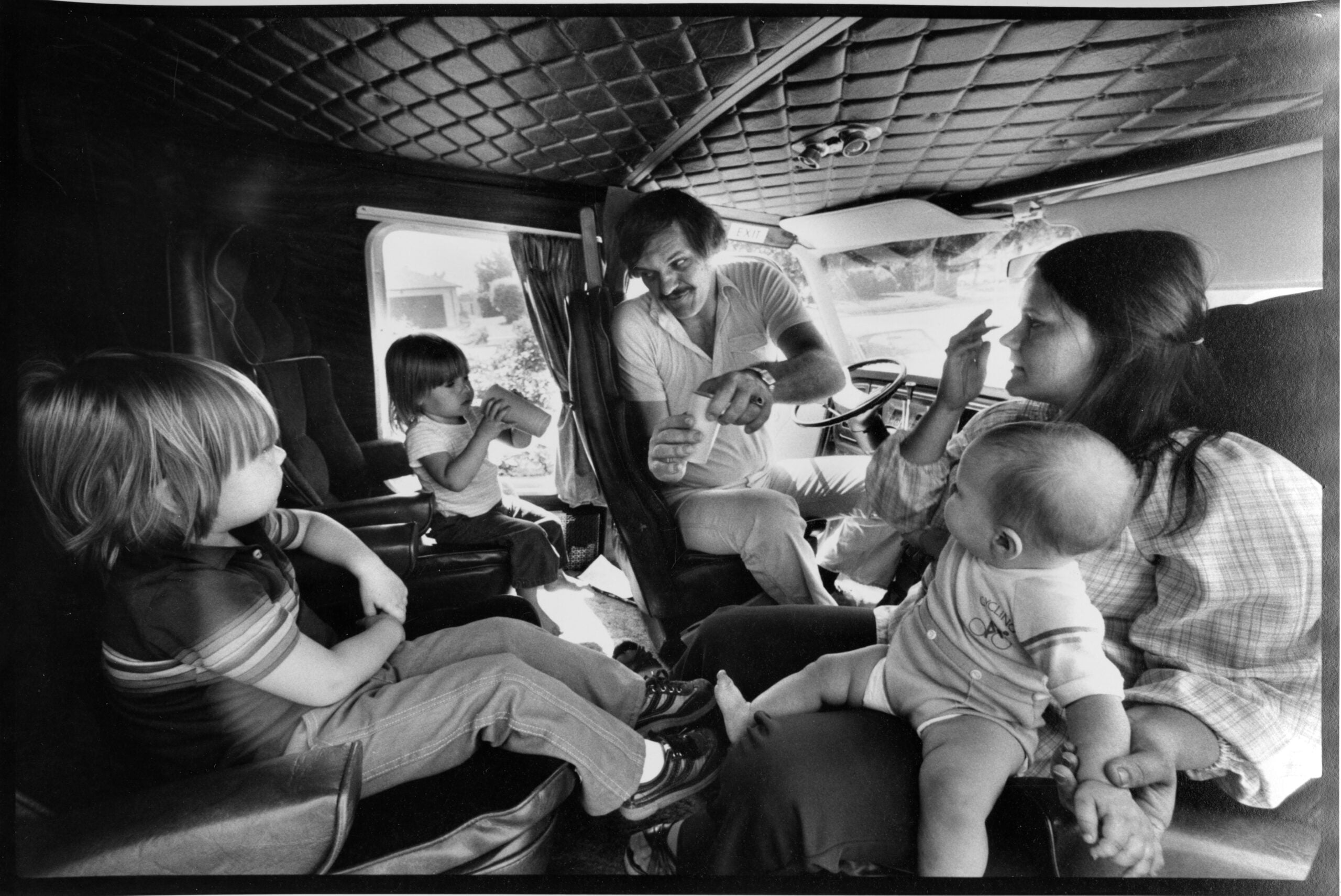 A man and woman sit in a vehicle with three young children. The woman holds a baby, while two kids sit nearby. The man appears to be talking to the family, gesturing with his hands.