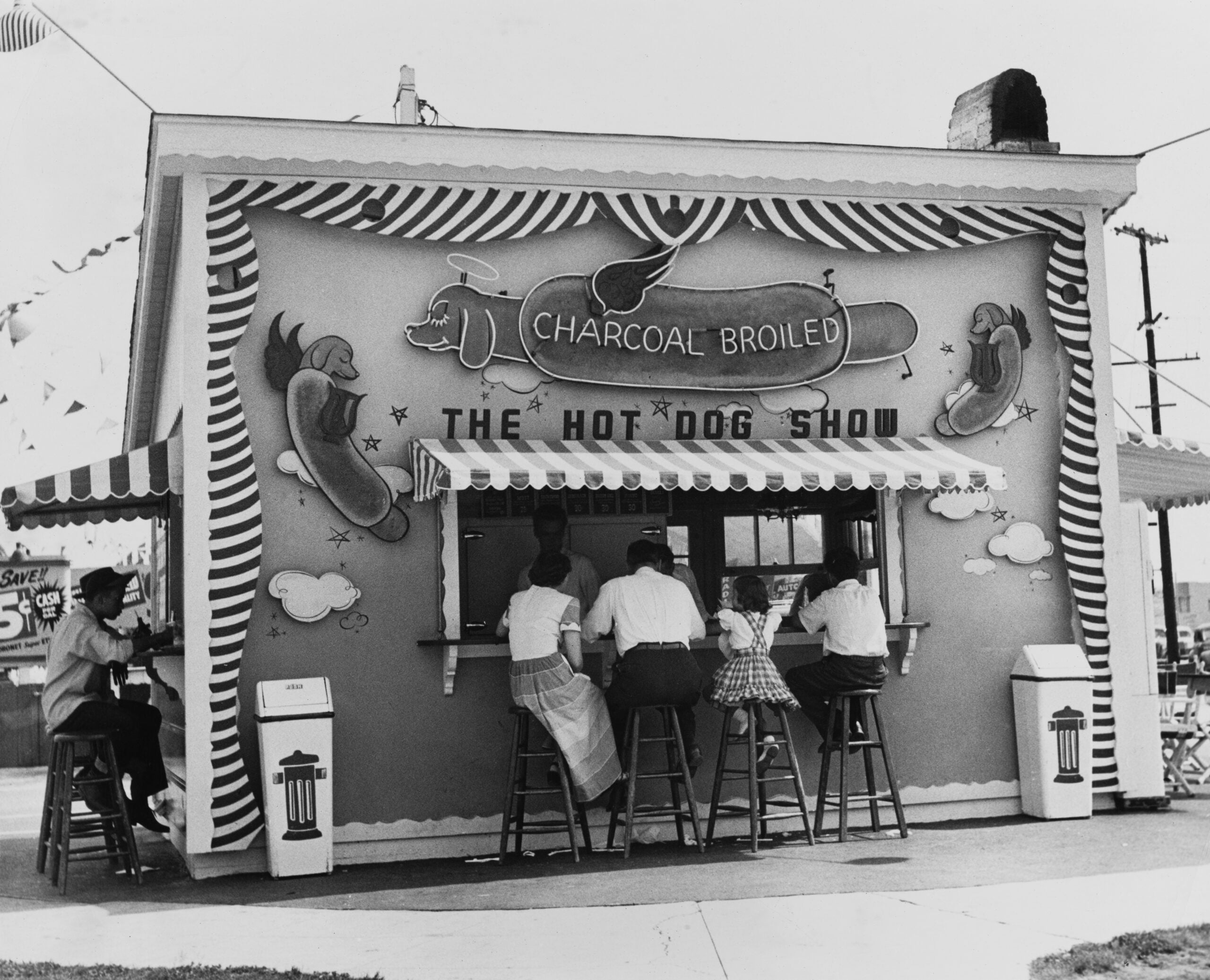 A black-and-white photo of people sitting at a whimsical hot dog stand with a striped awning, large hot dog sign, and playful decorations. The stand advertises “Charcoal Broiled” hot dogs at “The Hot Dog Show.”