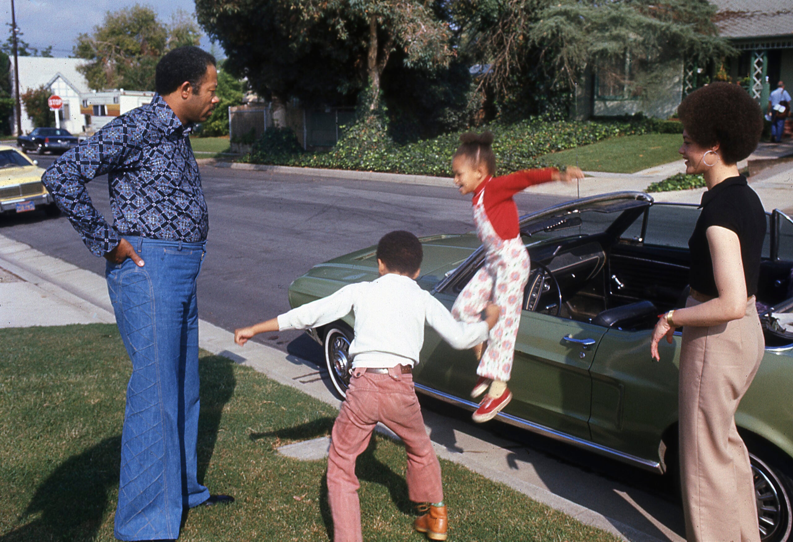 A man and woman watch as two children play energetically near a vintage green car parked on a suburban street. One child is jumping, while the other stands ready to catch them. Trees and houses are visible in the background.