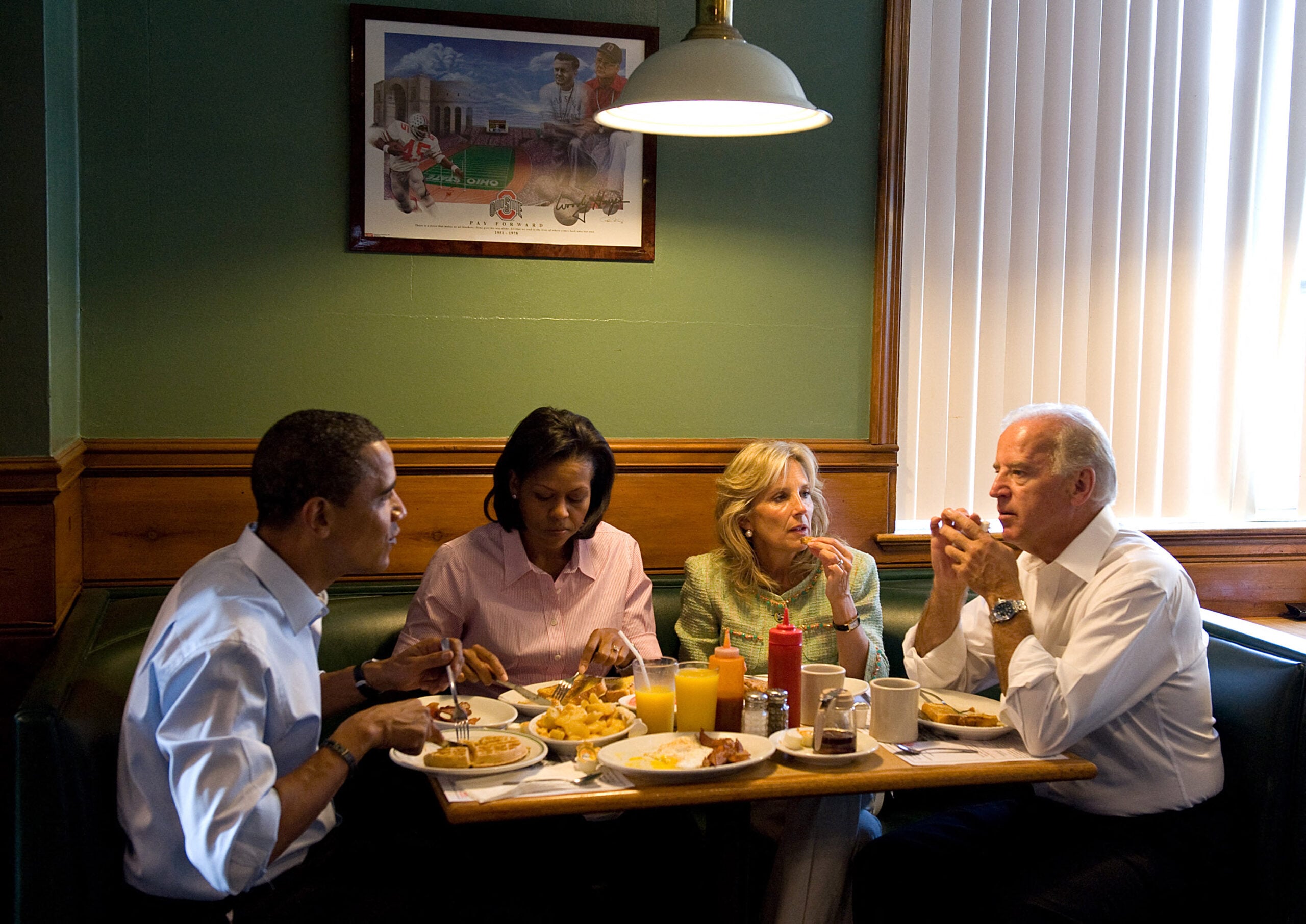 Four adults sit in a diner booth eating and talking. Plates of food, drinks, and condiments are on the table. A framed picture hangs on the green wall behind them. Light streams in from a window with blinds.