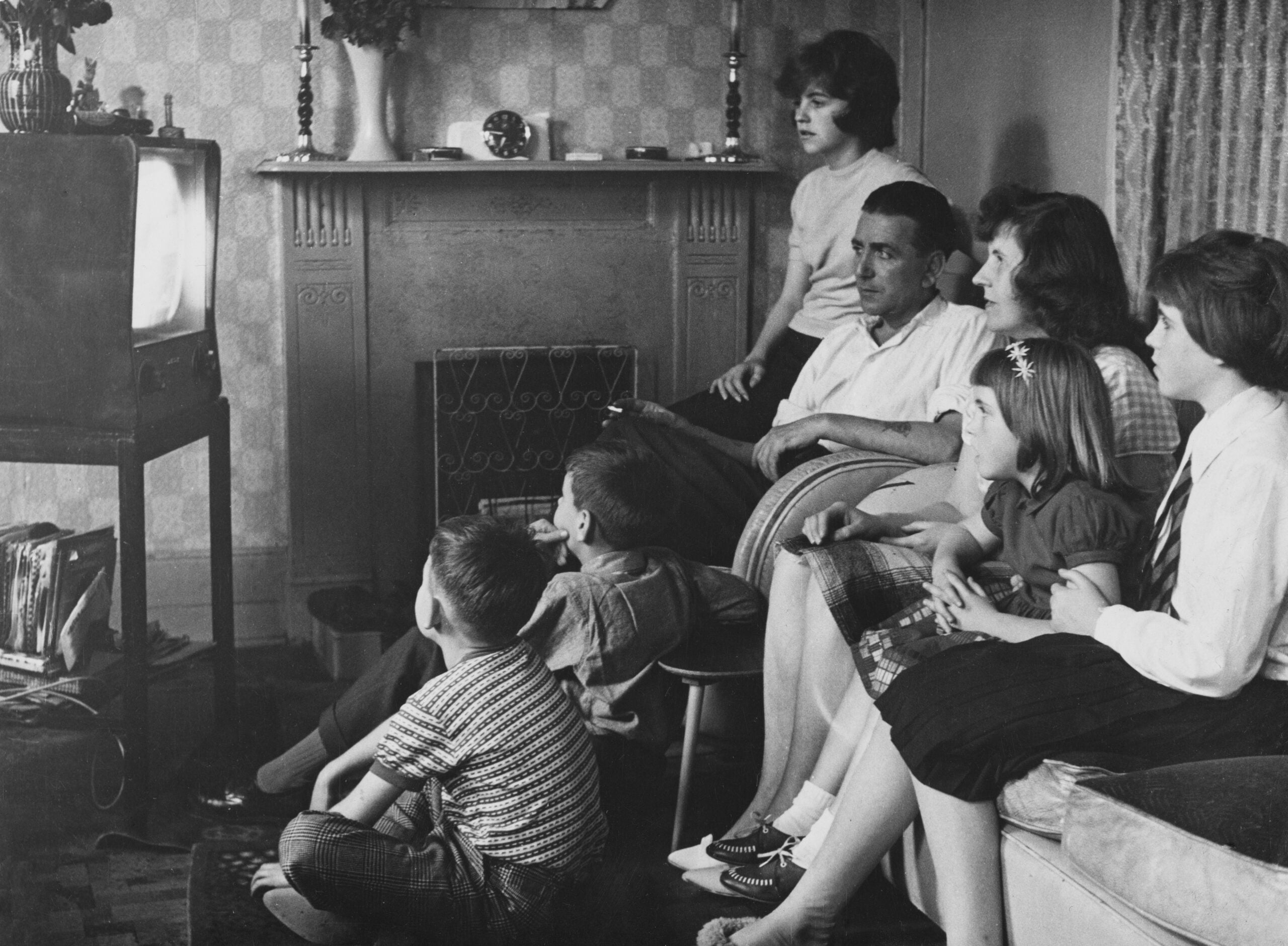 A family of seven sits together in a living room, watching a black-and-white television. Some are on the sofa, while children sit on the floor. The scene appears to be from the mid-20th century.