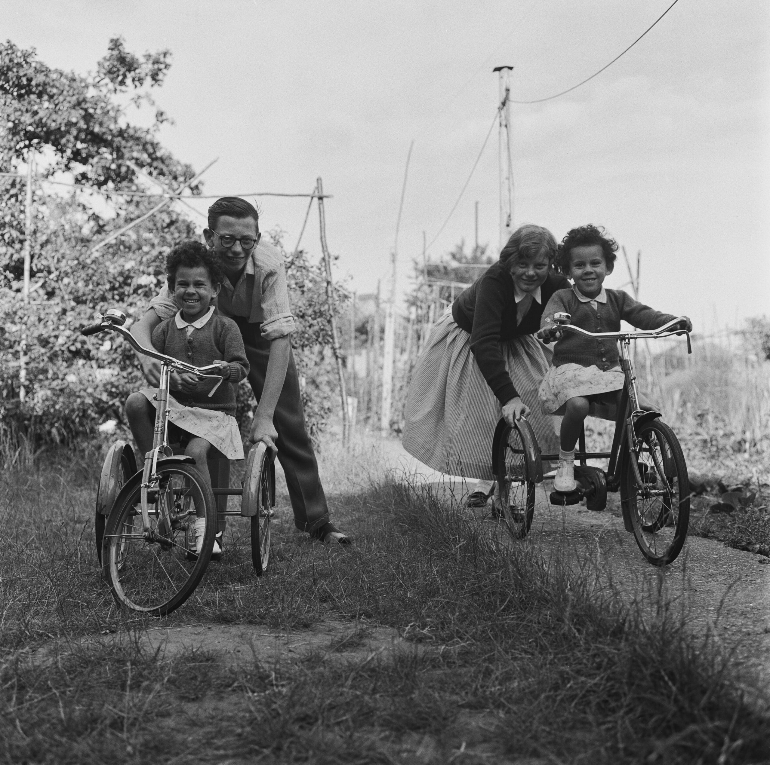 Two adults help two smiling children ride tricycles on a path in a grassy backyard, surrounded by trees and garden poles, under a partly cloudy sky.
