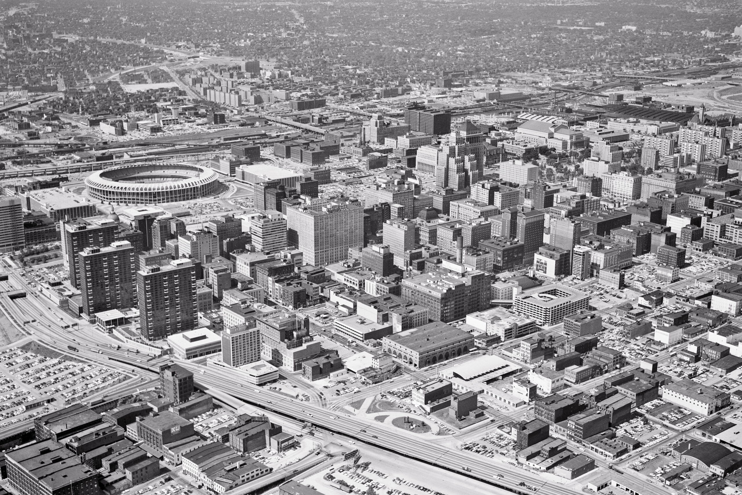 Aerial black and white view of a dense city with many buildings, roads, and a large round stadium near the center. The scene includes cars, intersections, and sprawling urban development.