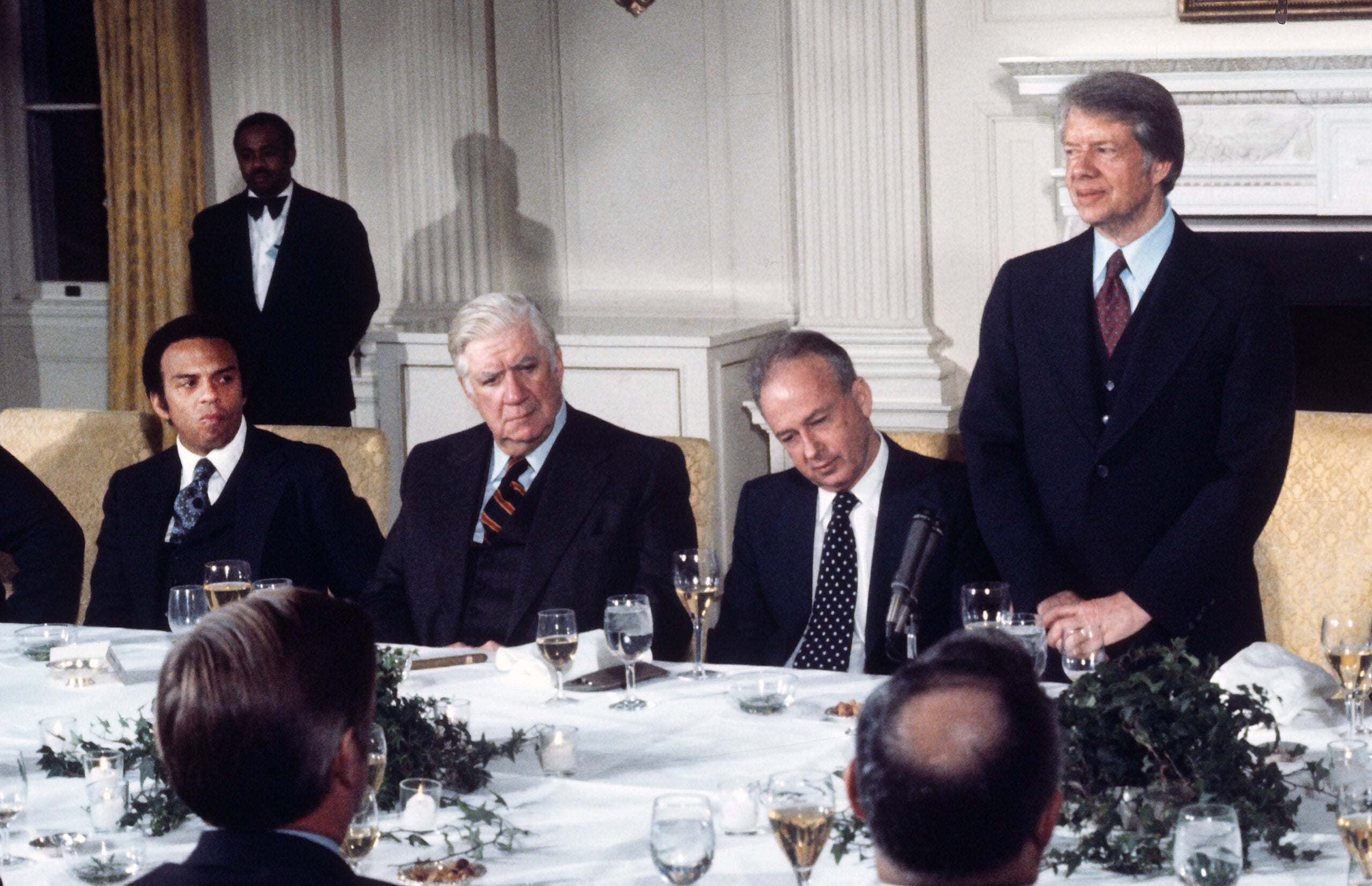 Four men in suits sit at a formal dining table, one man standing and speaking while others listen. Glasses, plates, and greenery are on the table in an elegantly decorated room.