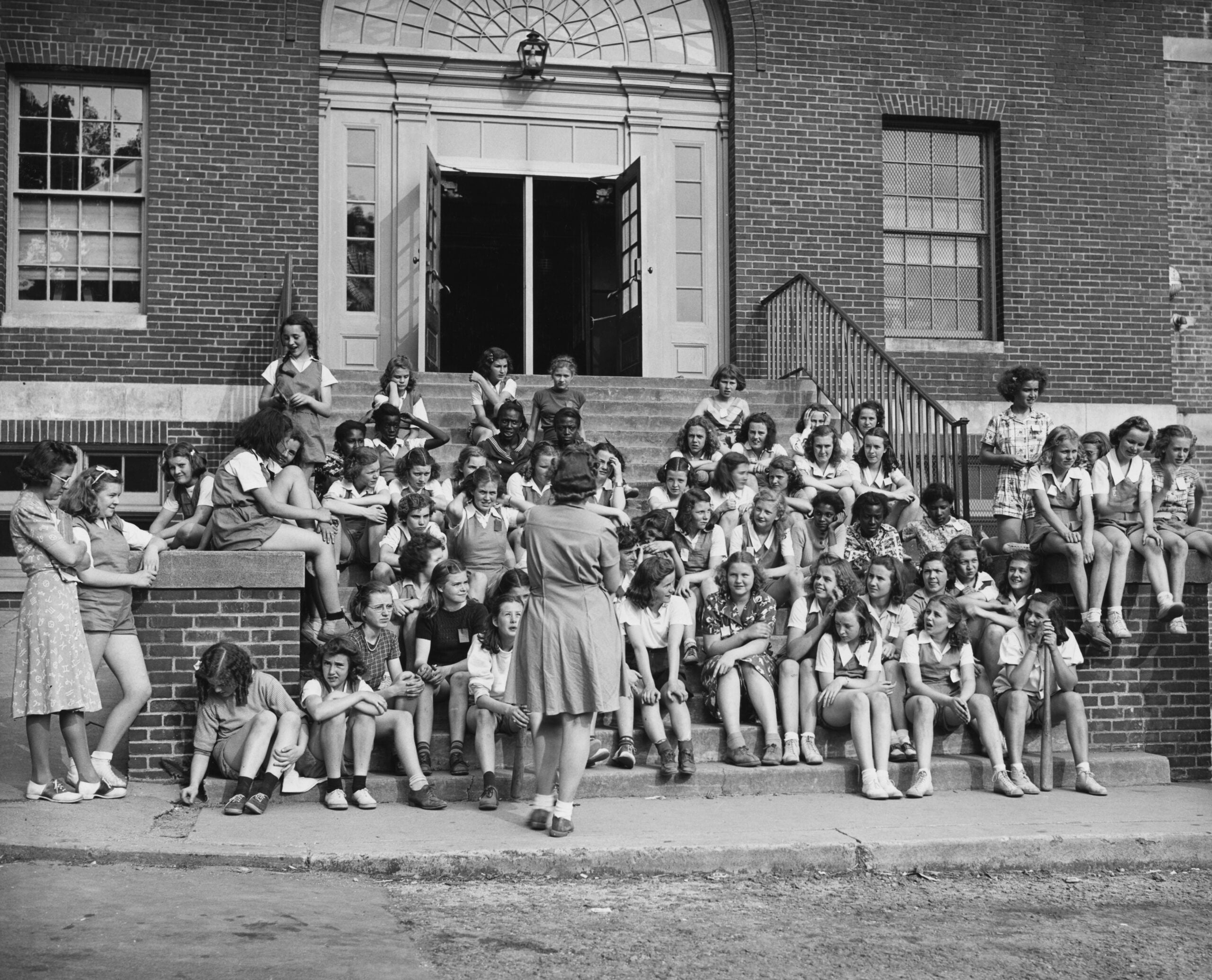 A large group of children sits and stands on the steps of a brick school building, listening to an adult woman who faces them with her back to the camera. The scene captures a glimpse of school life in the 1960s and 70s, with the doors of the building open behind them.