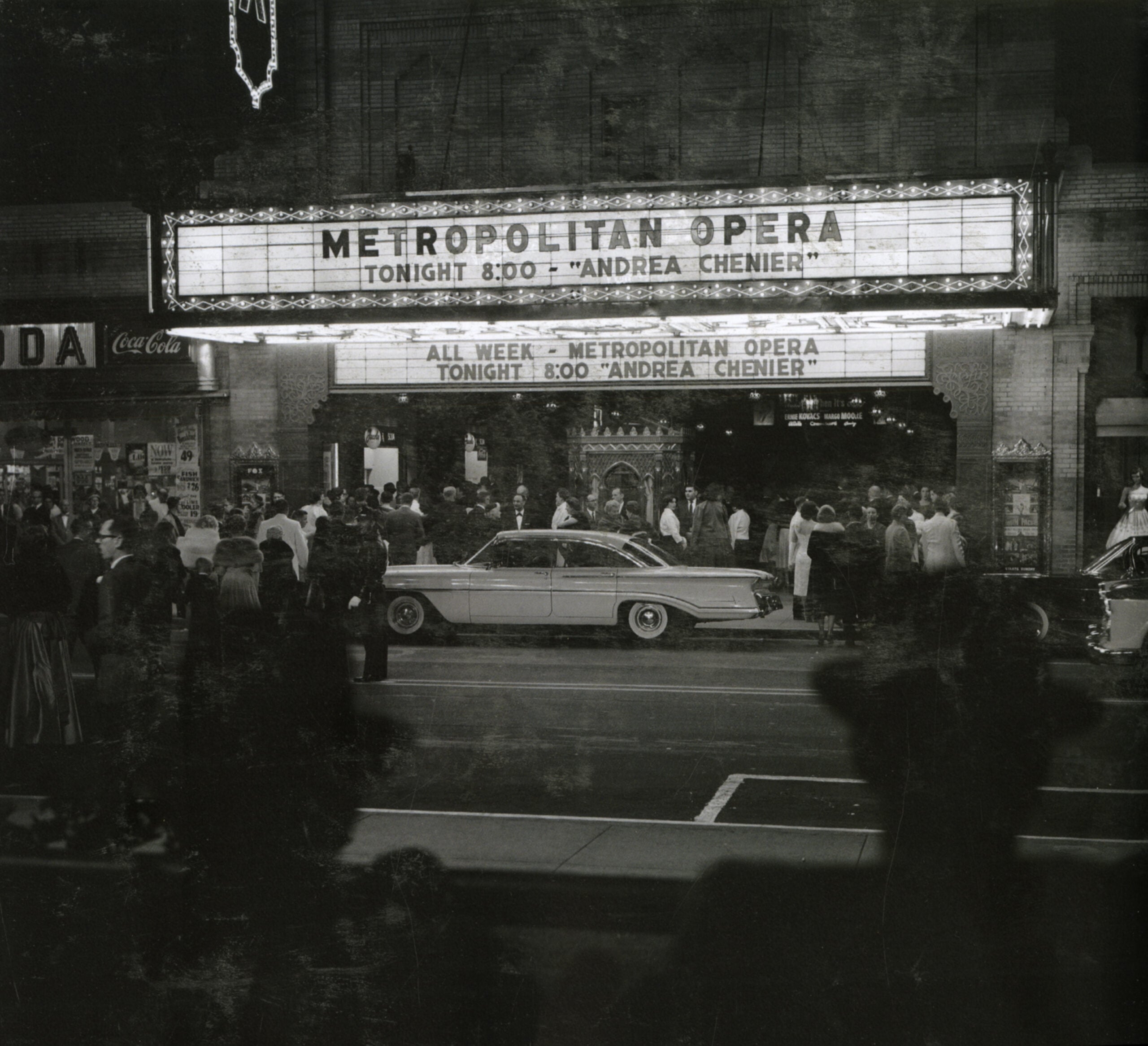 Crowd and vintage car outside Metropolitan Opera at night. Marquee reads: “Metropolitan Opera Tonight 8:00 ‘Andrea Chenier’” and advertises the show running all week. The scene is lively and illuminated.