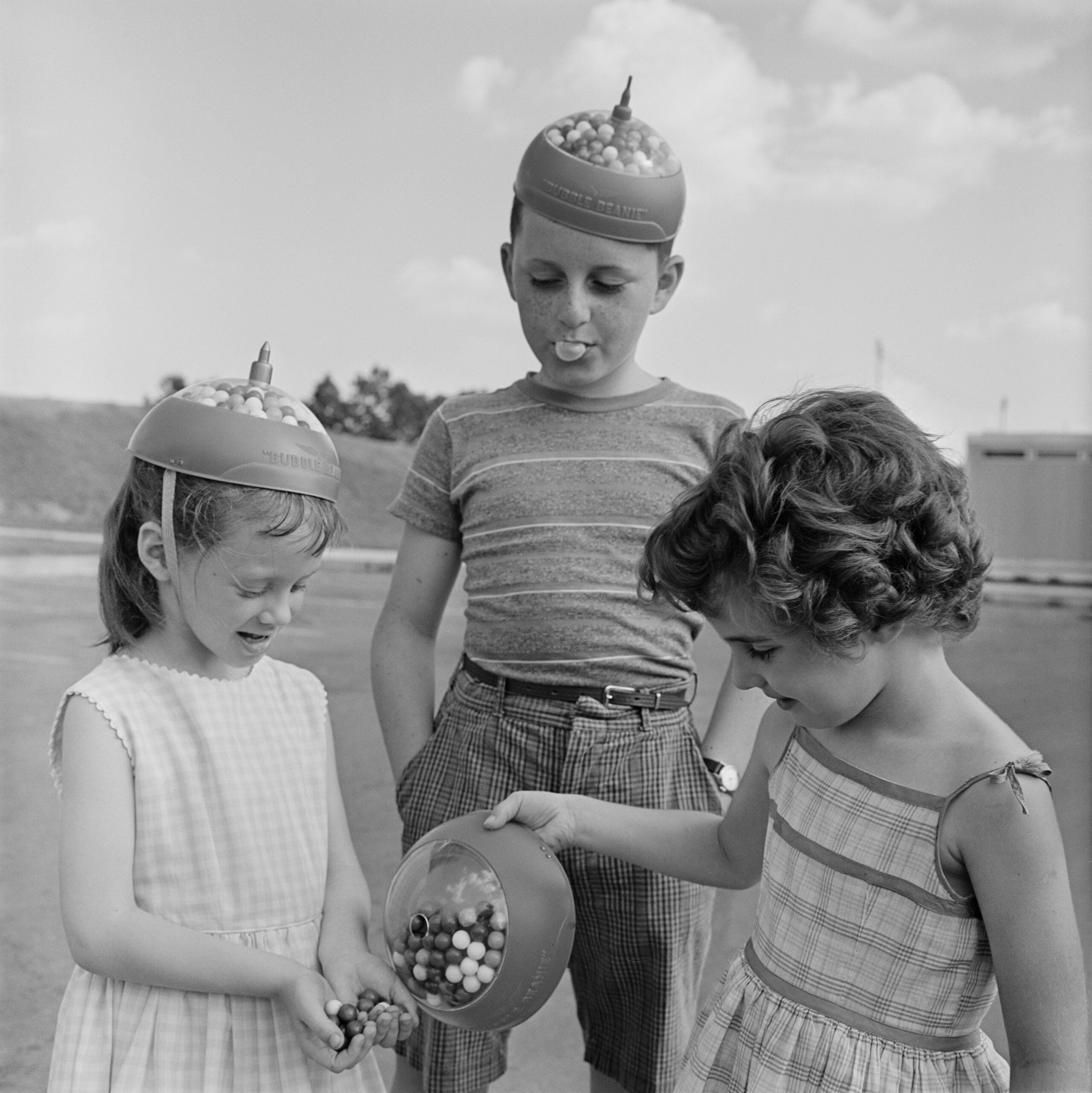 Three children wearing plastic hats filled with gumballs, with one girl handing gumballs to another while the boy blows a bubblegum bubble. They are outdoors under a partly cloudy sky.