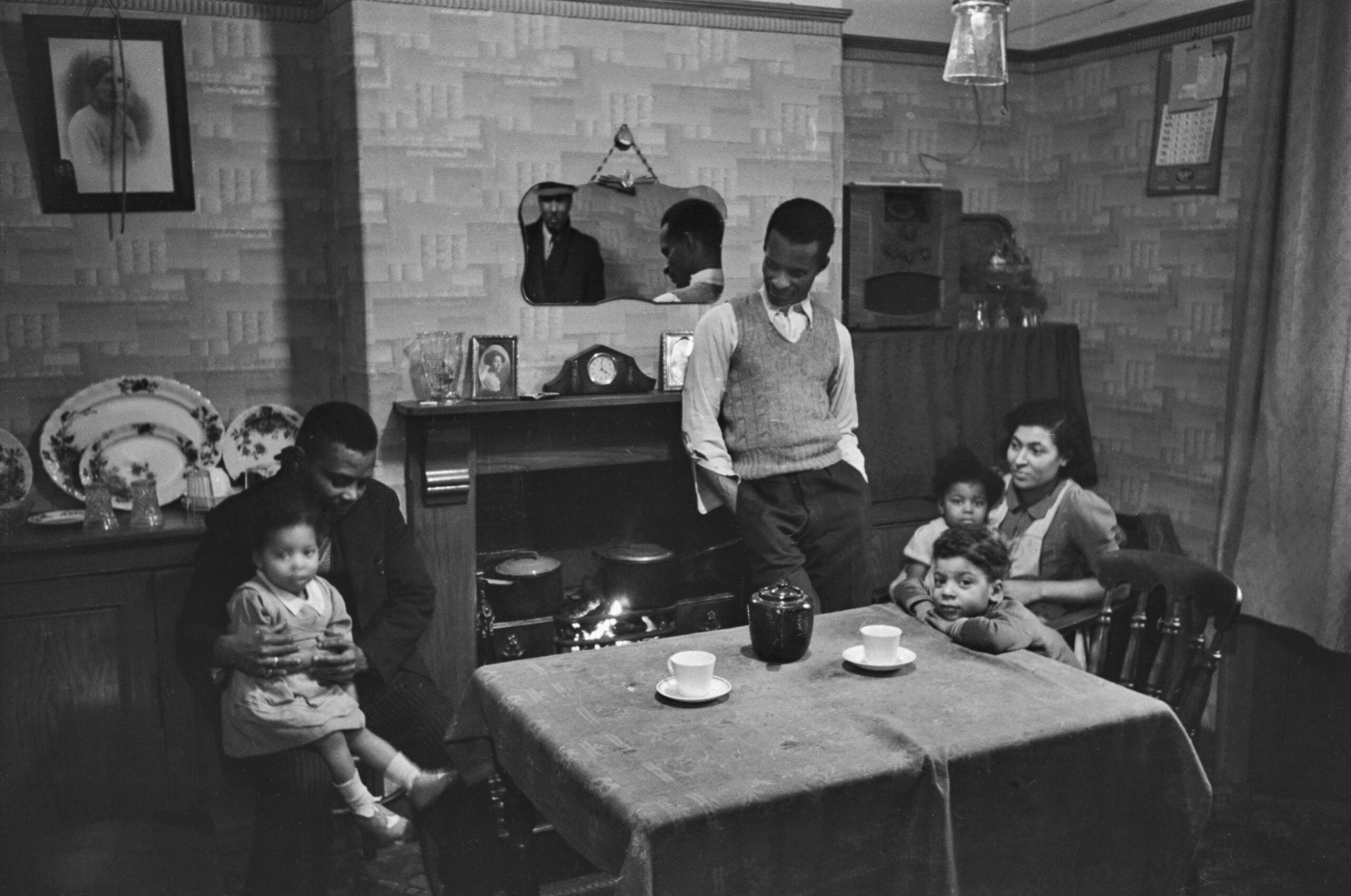A family of six sits and stands around a fireplace in a cozy, wallpapered living room with a set table for tea, vintage decor, and family portraits on the wall.